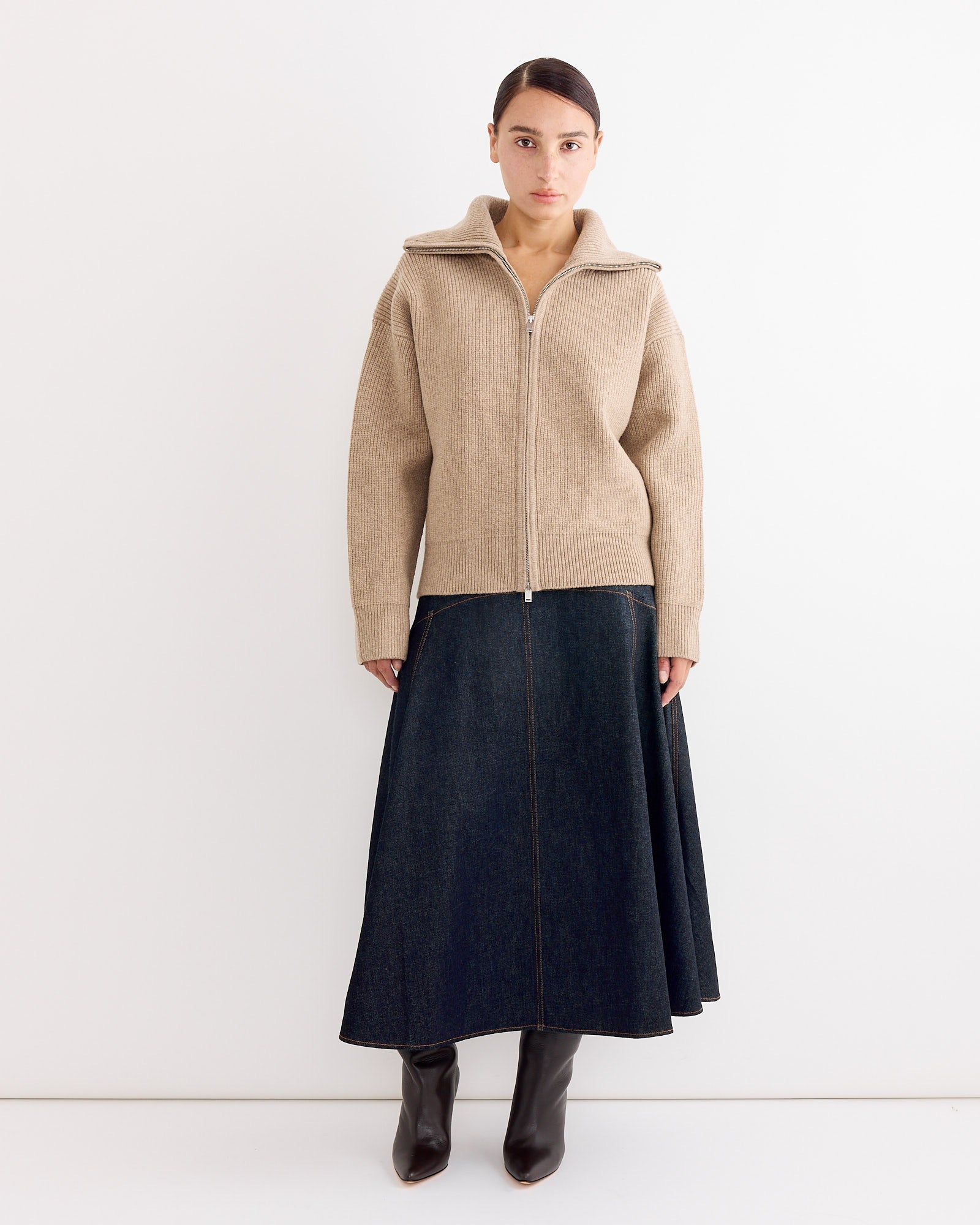A woman with brown hair pulled back wears the Studio Nicholson Sandos Jumper in Oatmeal, made from extrafine merino wool, styled with a long dark denim skirt and dark brown heeled boots against a white background.