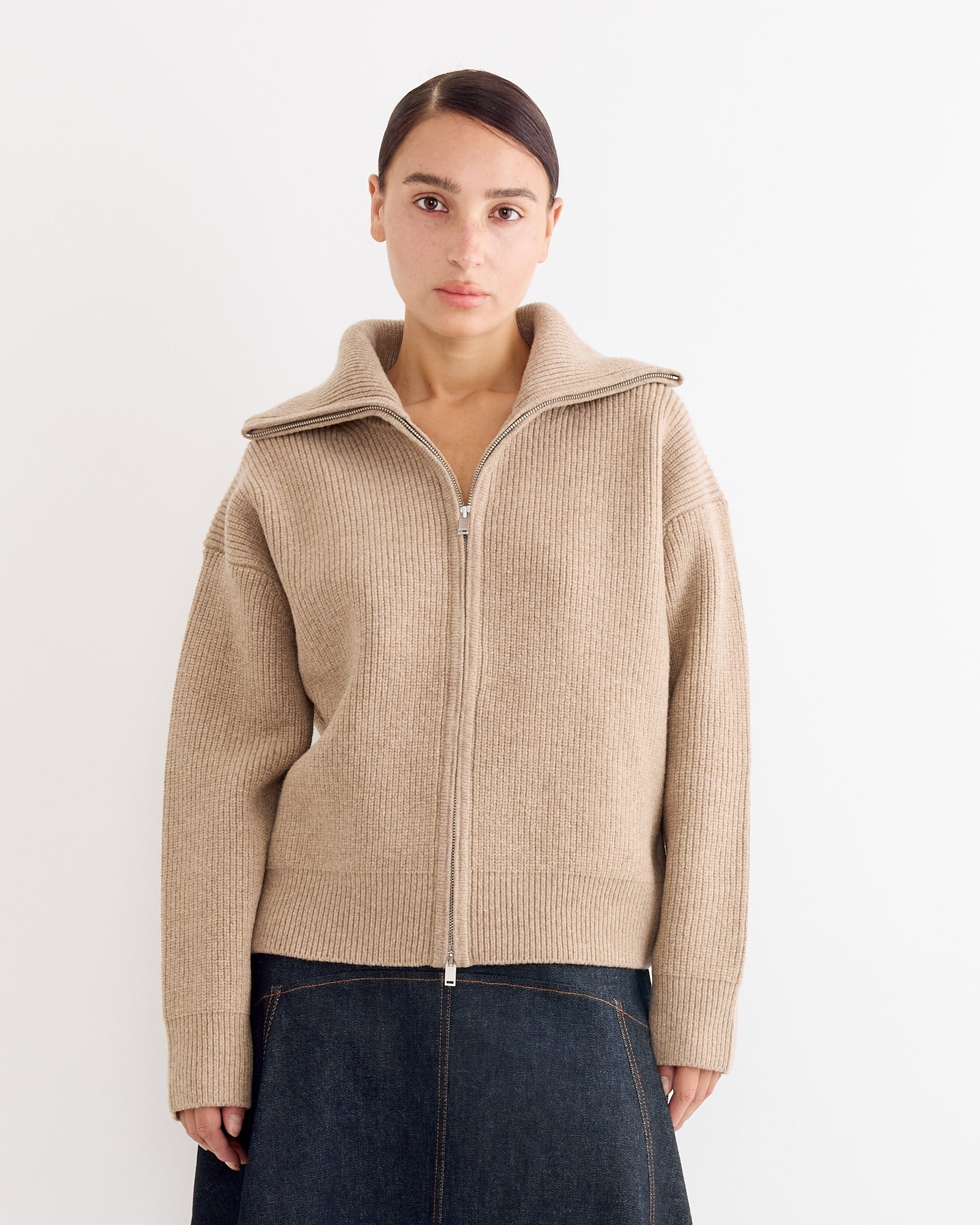 A person with straight brown hair pulled back wears the Studio Nicholson Sandos Jumper in Oatmeal, featuring extrafine merino wool and a wide collar, paired with a dark denim skirt, standing against a plain white background.