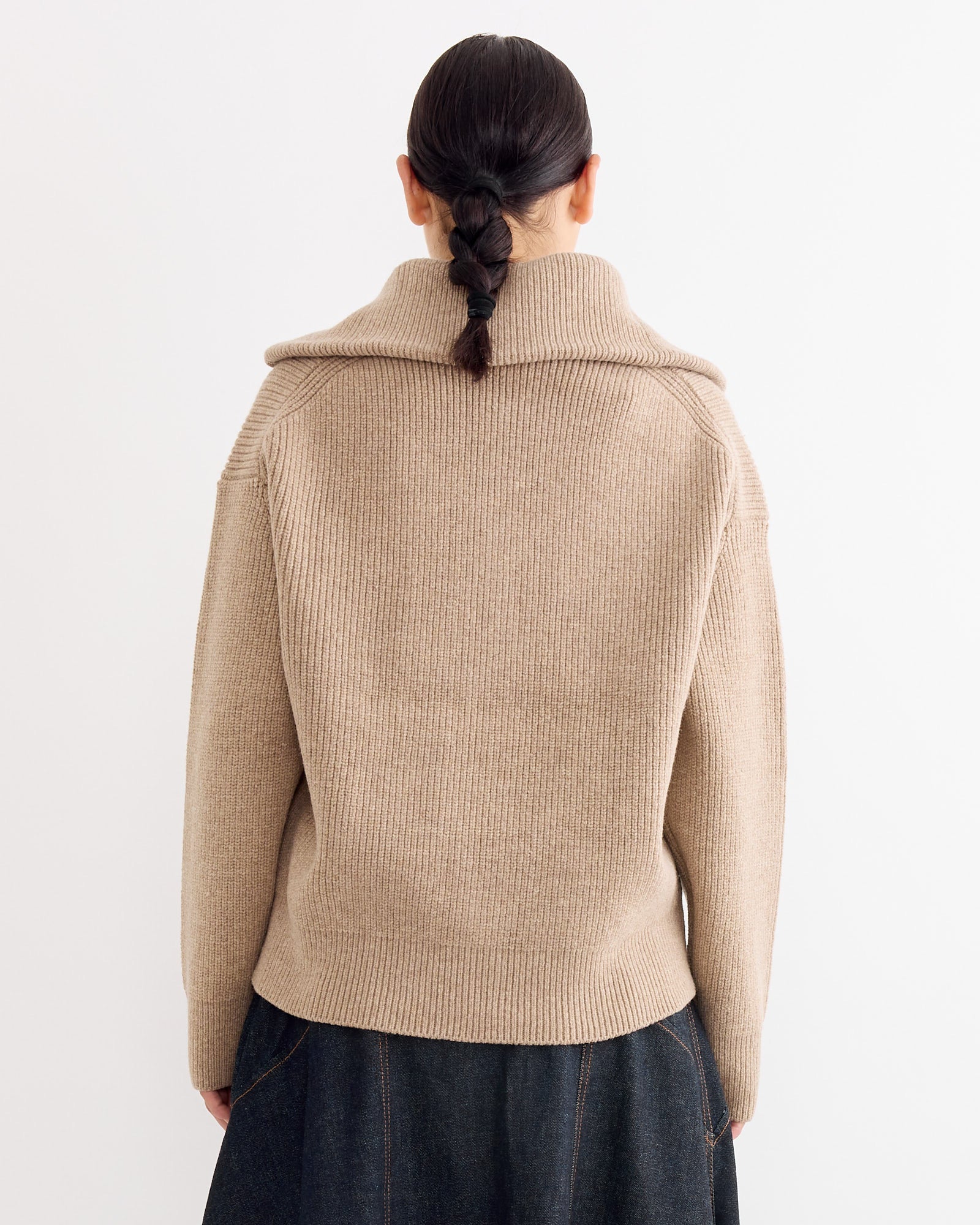 A person with dark braided hair is shown from the back, wearing the Studio Nicholson Sandos Jumper in Oatmeal with a wide collar and a dark skirt, standing against a plain white background.