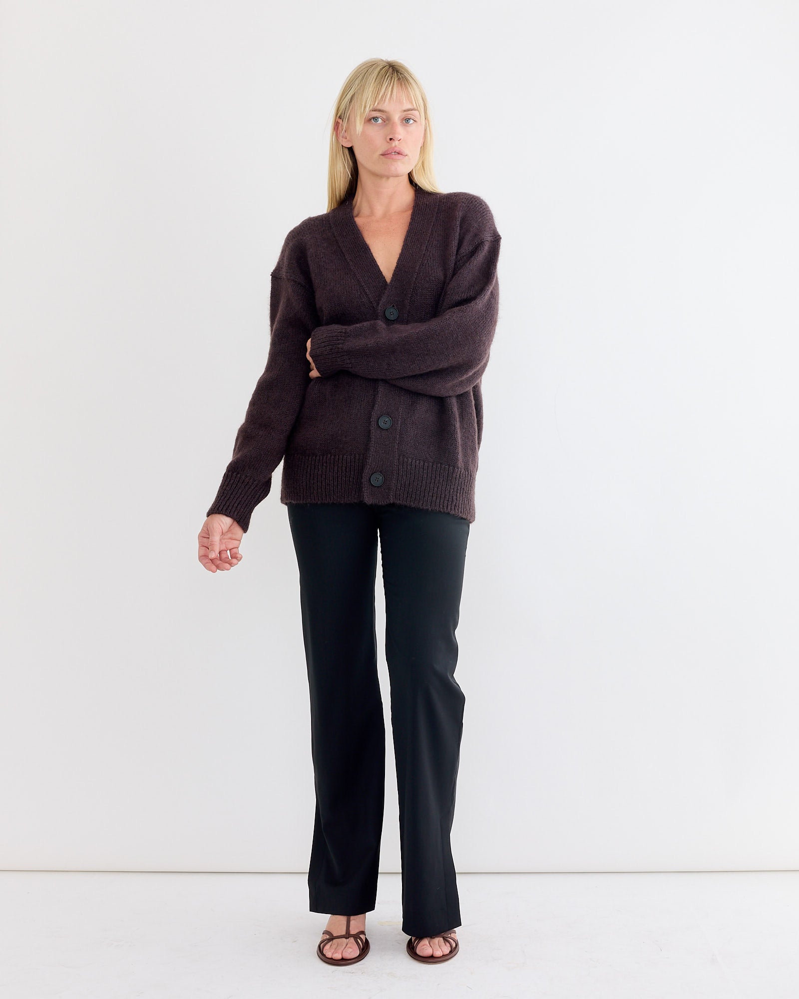 A woman with blonde hair and a neutral expression stands against a white wall wearing the Studio Nicholson Trey Cardigan in Espresso, black pants, and black sandals. One arm is relaxed while the other is slightly bent.