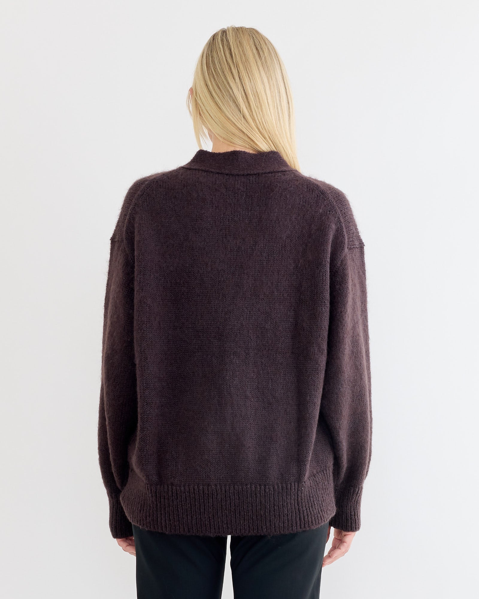 A person with long blonde hair, seen from behind, wears a loose-fitting Trey Cardigan in Espresso by Studio Nicholson and black pants against a plain white background.