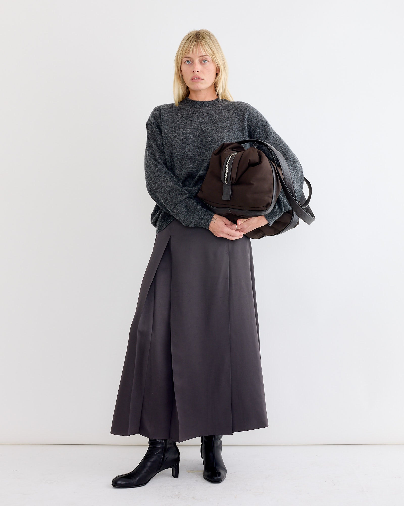 A blonde woman wears the Studio Nicholson Picco Crewneck in Charcoal with a long dark skirt, black ankle boots, and carries a large brown bag, standing against a white background.