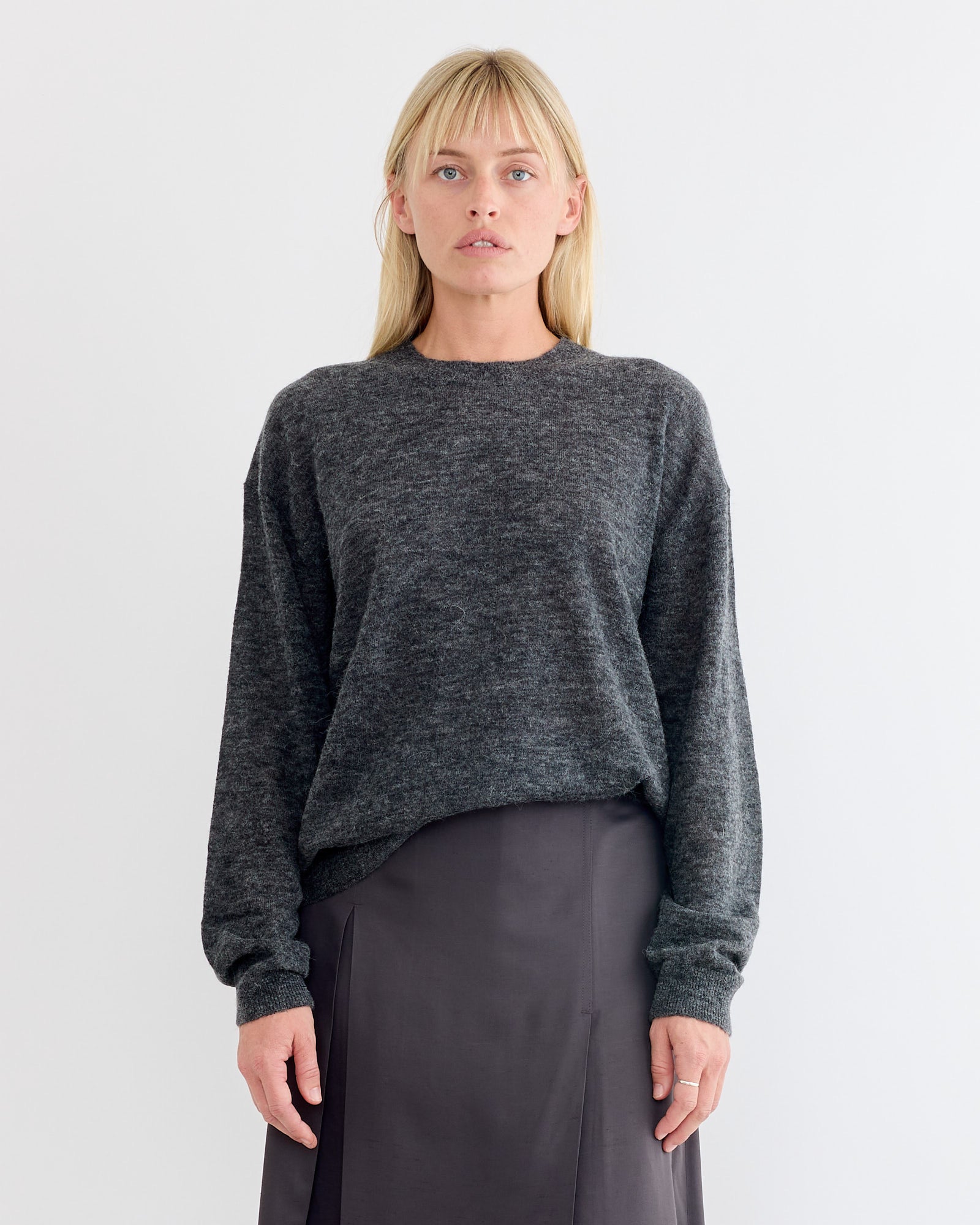 A woman with straight blonde hair and bangs wears the Studio Nicholson Picco Crewneck in Charcoal, paired with a charcoal skirt. She stands against a plain white background, looking directly at the camera with a neutral expression.