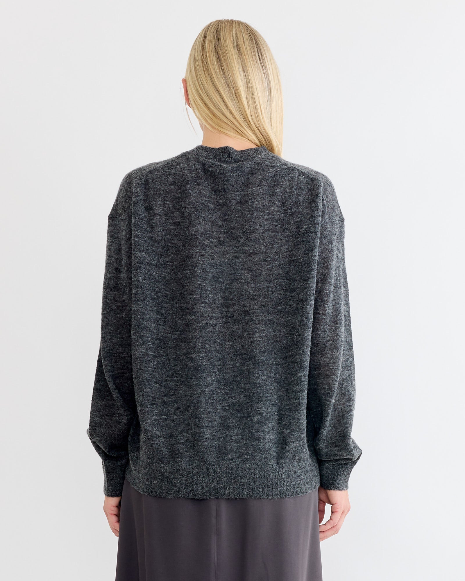 A person with long blonde hair, seen from behind, is wearing the Studio Nicholson Picco Crewneck in Charcoal and a dark skirt against a plain light background.