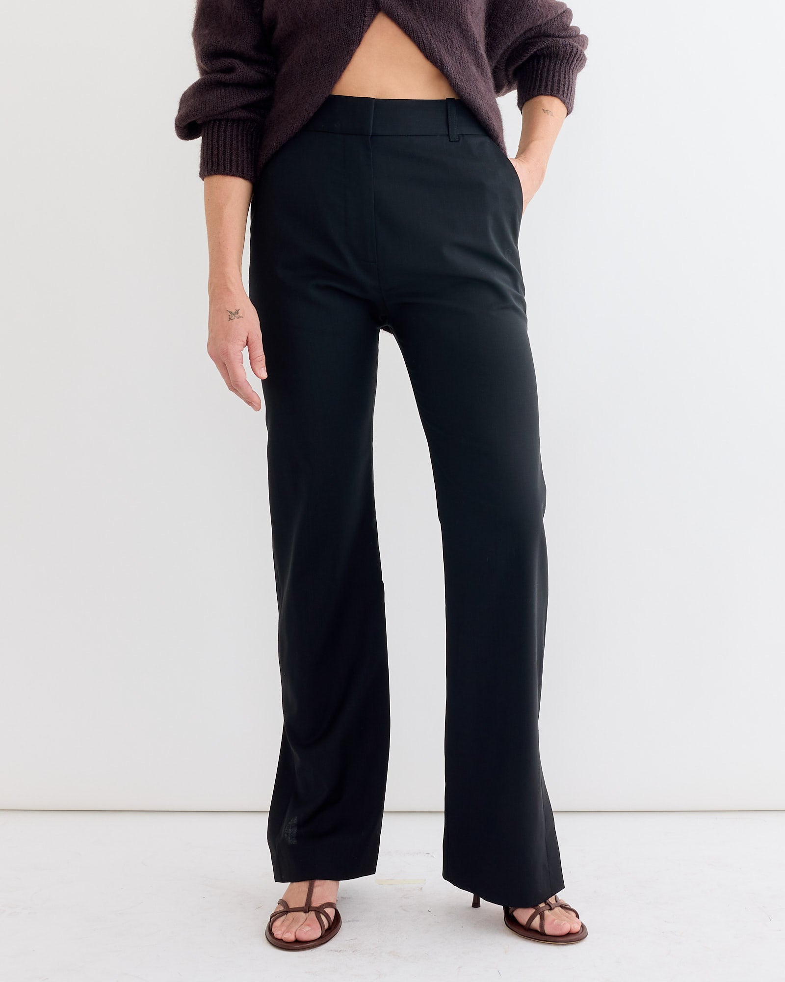 A person stands against a white background wearing Studio Nicholson's Mitra Pant in Black—tailored, high-waisted, wide-leg trousers—paired with a dark sweater, brown sandals, one hand in pocket, and midriff partially visible.