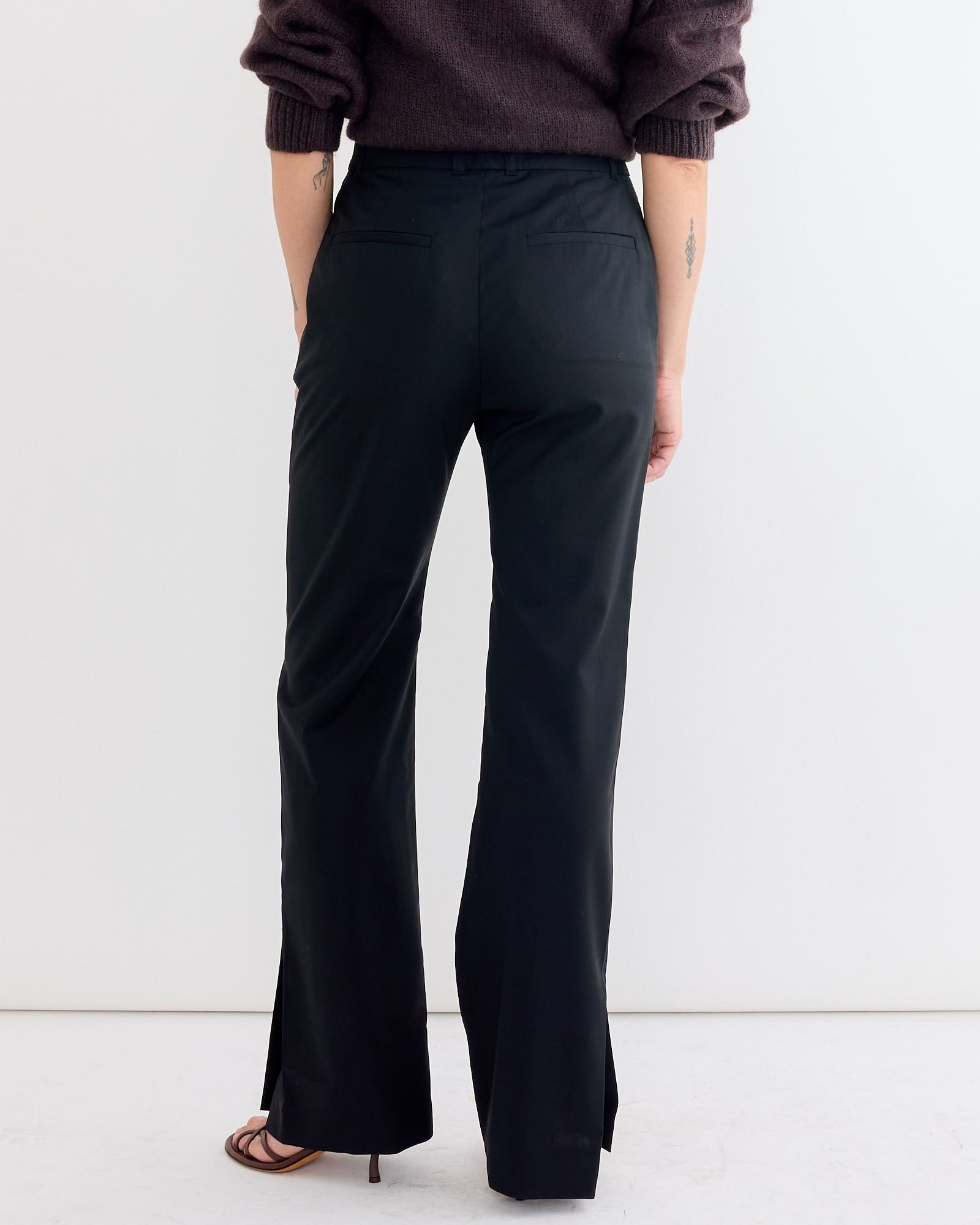 A person stands facing away against a white background, wearing the Studio Nicholson Mitra Pant in Black with hem slits, paired with a dark long-sleeve top and open-toe heeled sandals.
