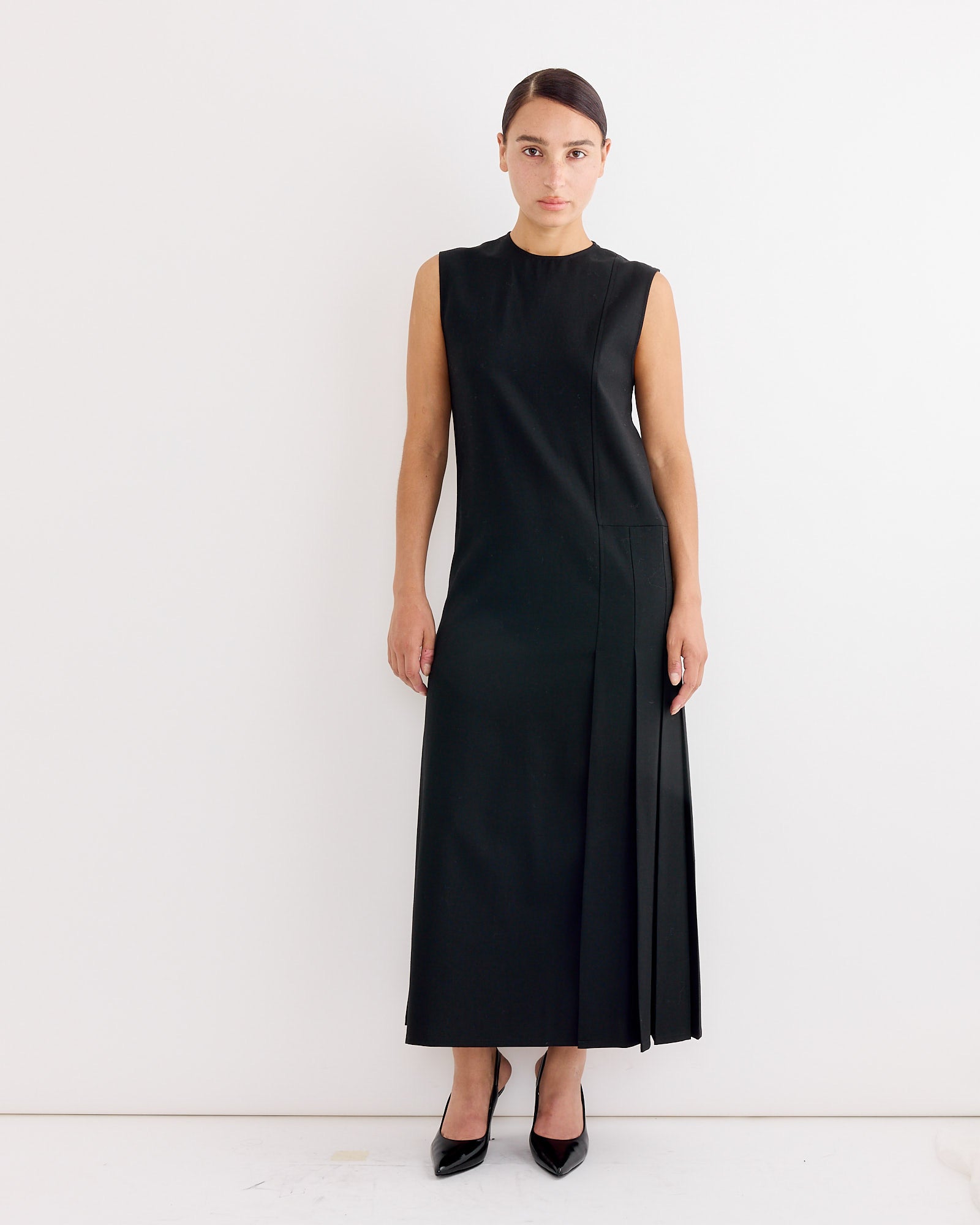 A woman with dark hair pulled back stands against a white wall wearing the Studio Nicholson Carat Dress in Black, a sleeveless, ankle-length pleated A-line dress, paired with black heels and her arms relaxed at her sides.