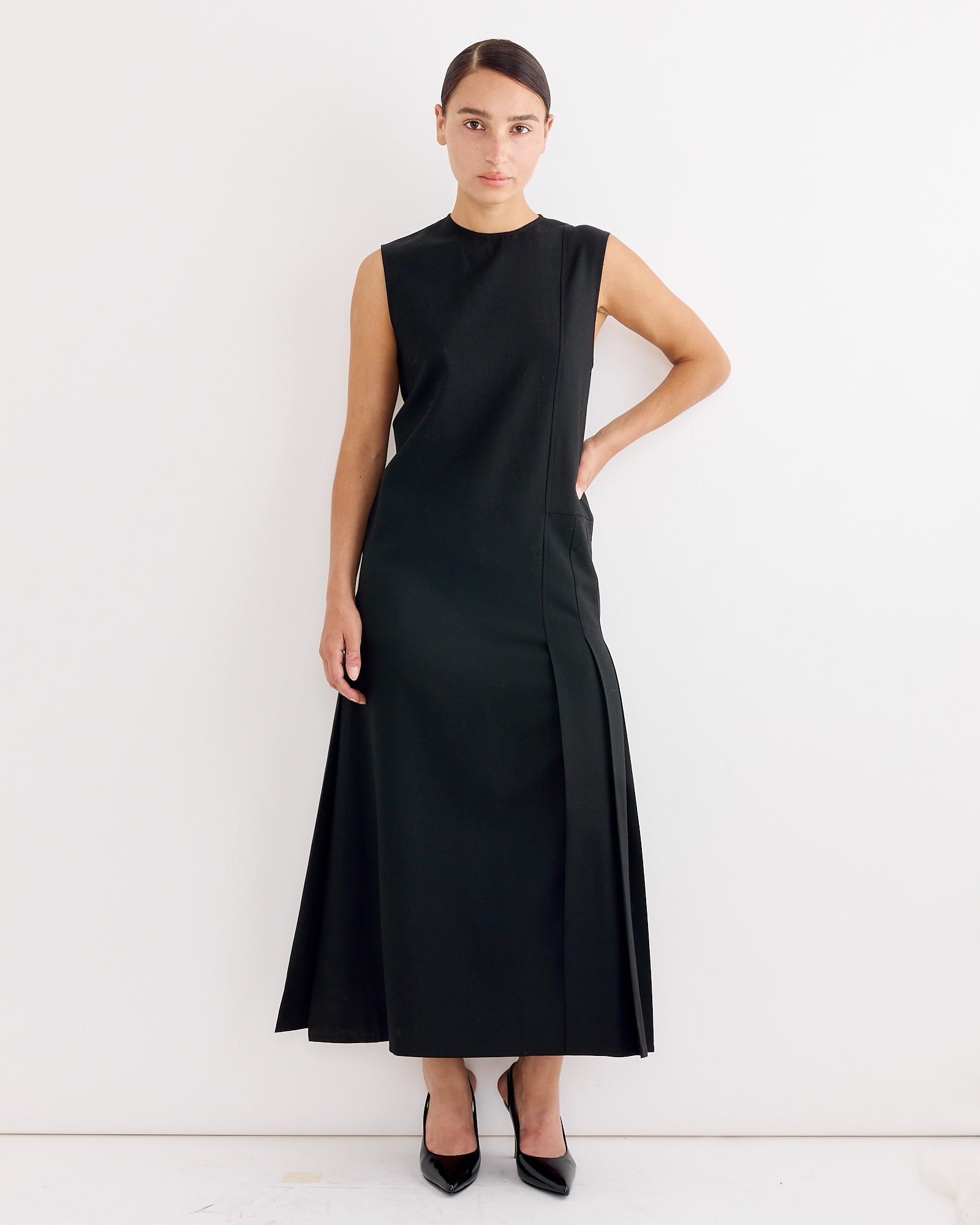 A woman stands against a white wall wearing the Studio Nicholson Carat Dress in Black with black heels. Her hair is neatly parted and pulled back, and she poses with one hand on her hip.