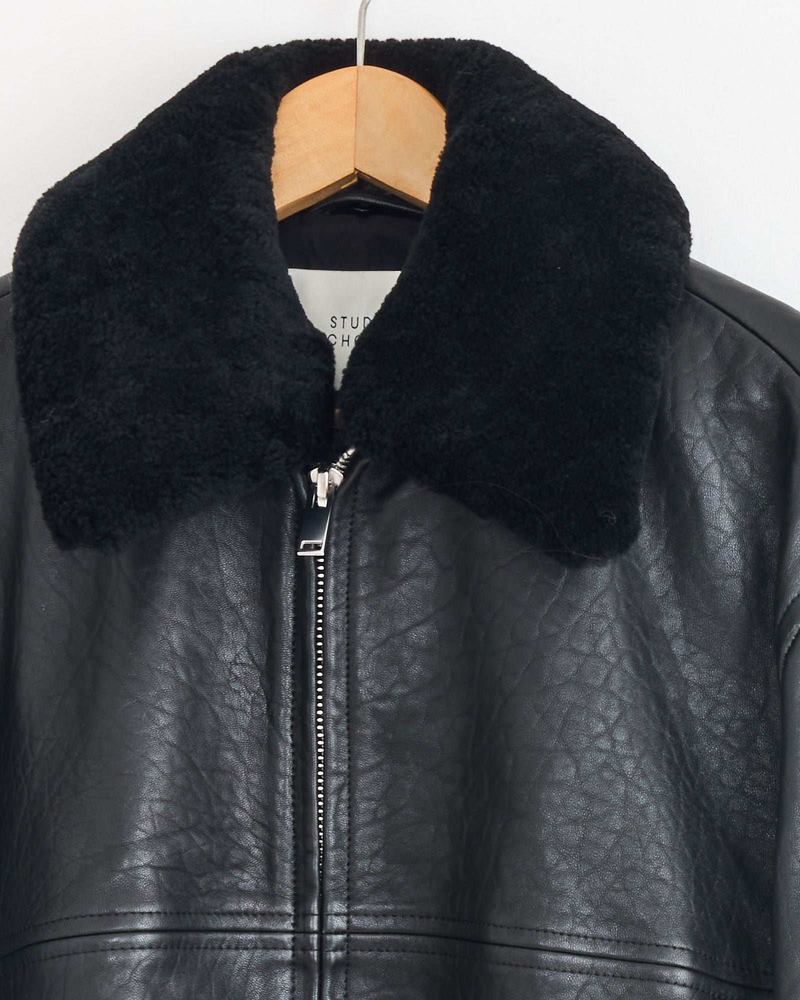 The Studio Nicholson Erie Jacket in Black, made from sheepskin leather with a large detachable shearling collar, hangs on a wooden hanger against a white background. It features a visible zipper and slightly wrinkled texture.