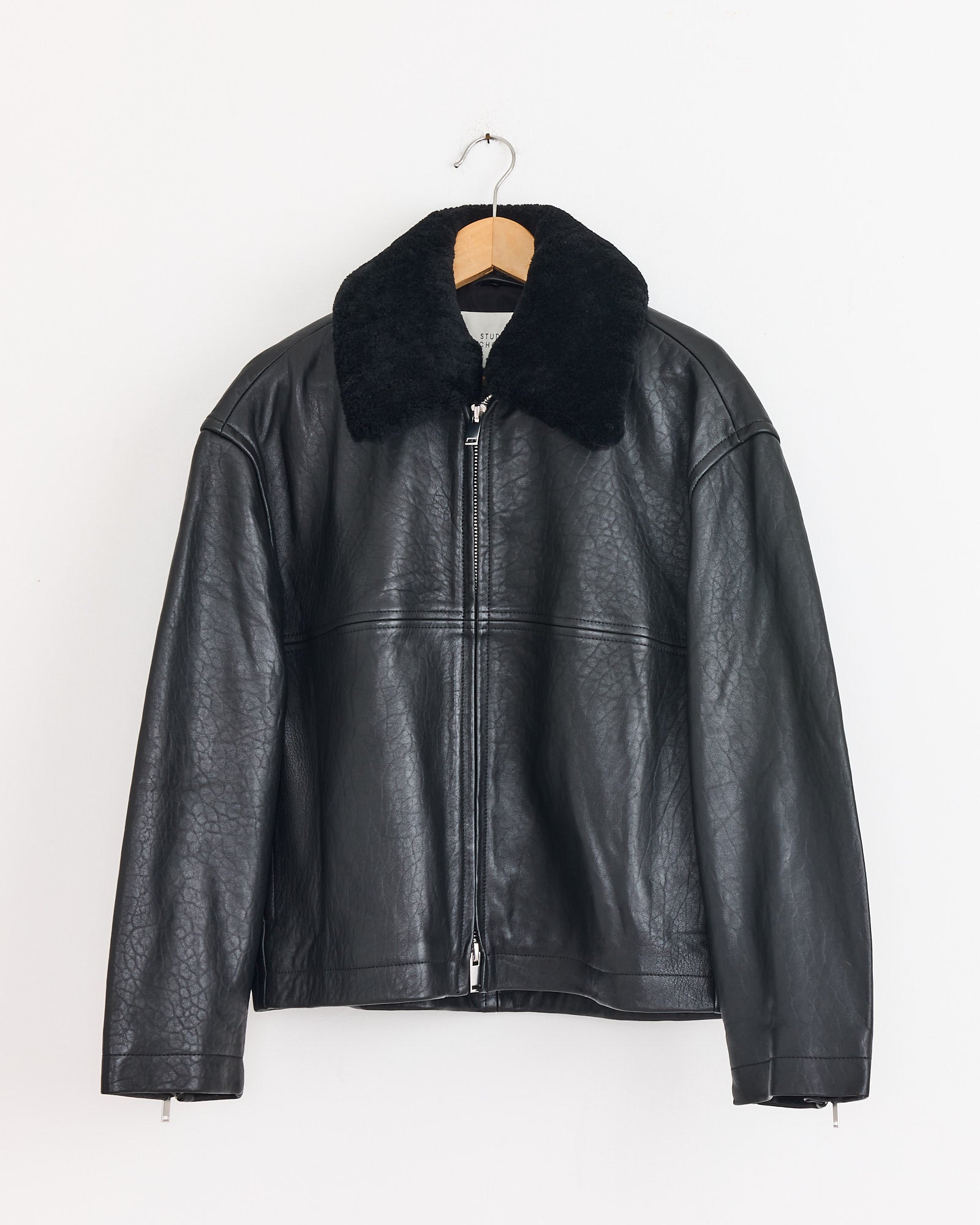 The Studio Nicholson Erie Jacket in Black, a sheepskin leather piece with a zipper front and oversized fur collar, is displayed on a wooden hanger against a plain white wall.