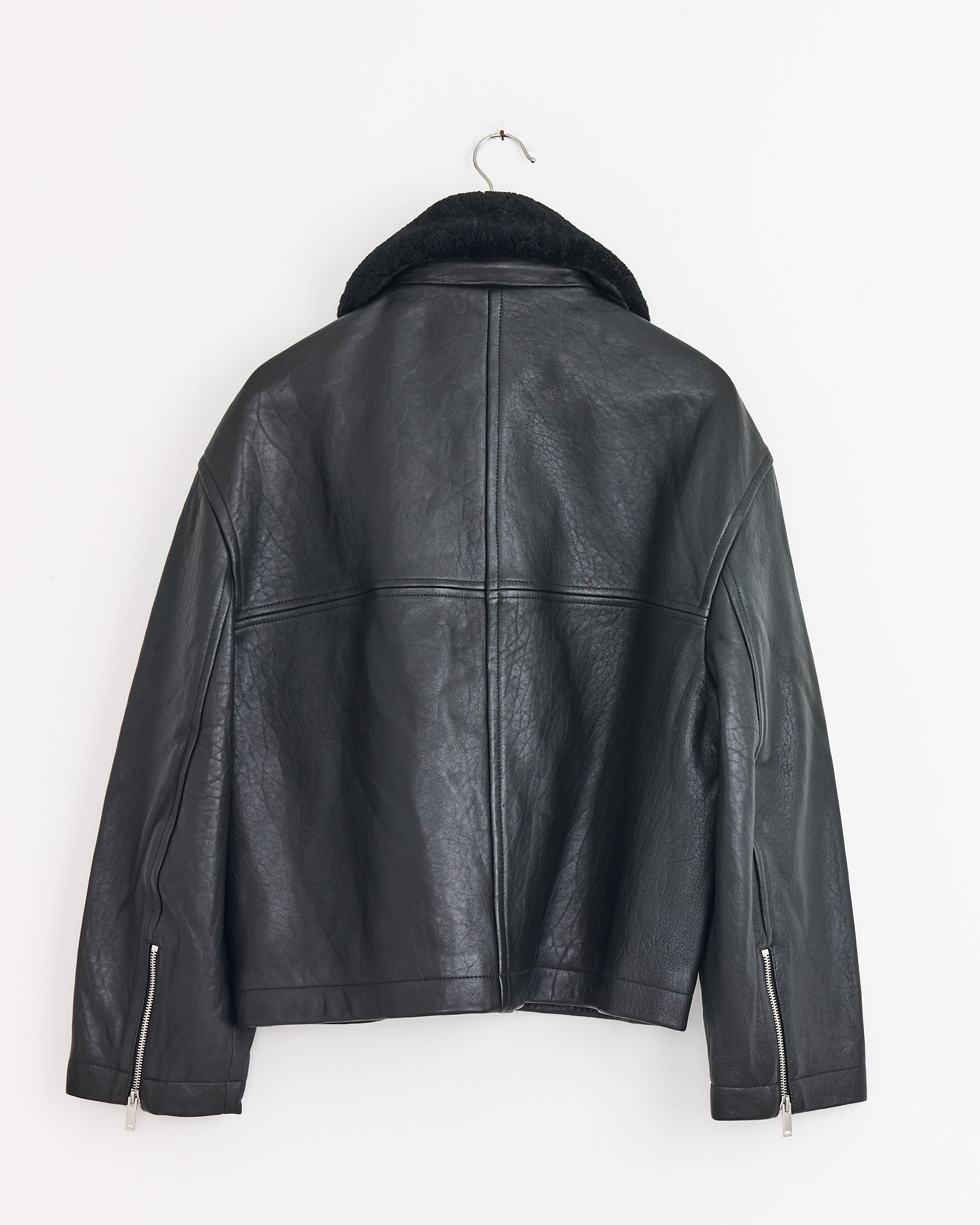 The Studio Nicholson Erie Jacket in Black, crafted from sheepskin leather with a detachable shearling collar, hangs on a hook against a white wall and features a textured finish plus zippered sleeve details.