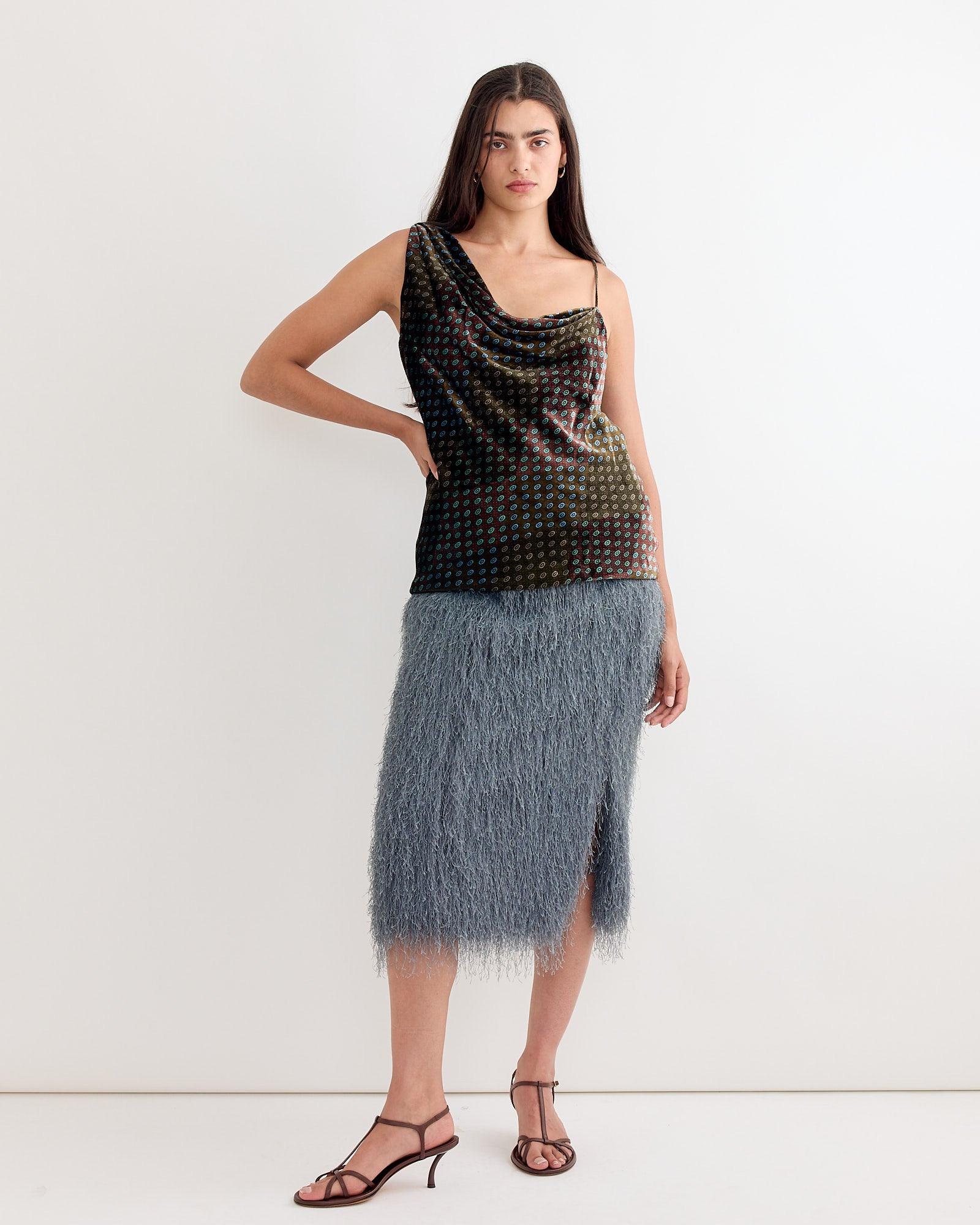 A woman with long hair stands against a white background, wearing Dries Van Notens Printed One Shoulder Top in Khaki and a textured, knee-length gray skirt with a front slit. She completes the look with strappy sandals.