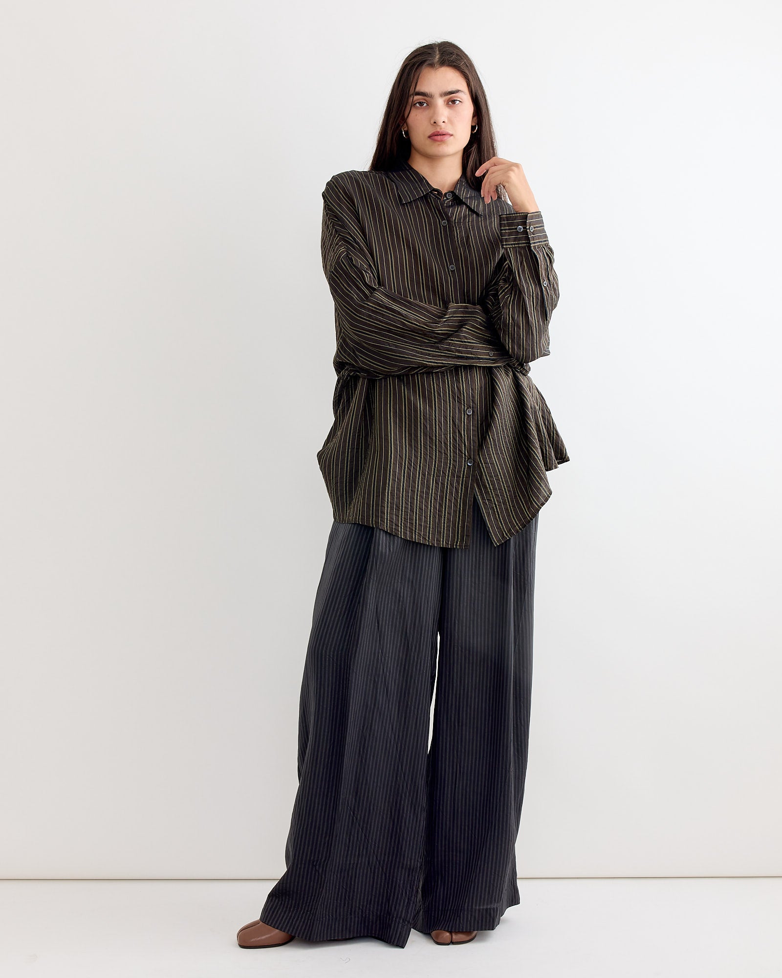 A person with long, dark hair and a neutral expression wears the Dries Van Noten Striped Shirt in Brown against a white background, paired with wide-leg dark pinstriped pants and brown shoes.