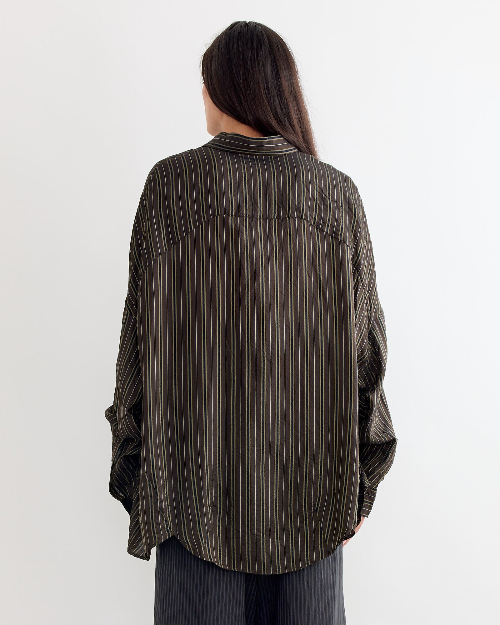 A person with long dark hair, seen from behind, wears the Dries Van Noten Striped Shirt in Brown with an oversized fit and loose dark pants against a plain light background.