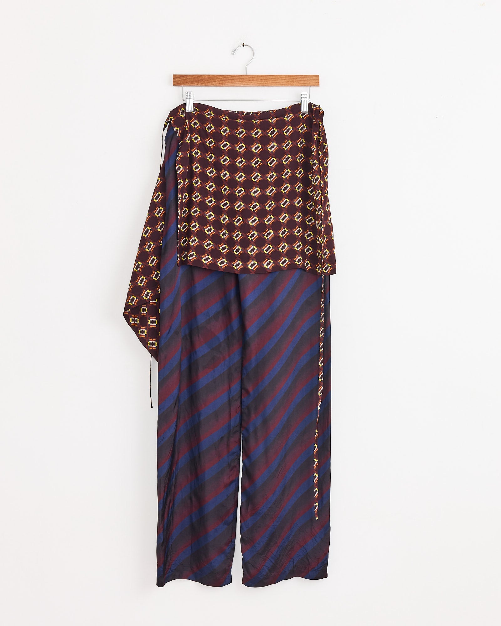 Dries Van Noten Printed Skirt Pants in Navy, featuring wide-leg stripes in navy and burgundy, with an attached asymmetrical maroon apron-like layer adorned with a yellow geometric pattern, displayed on a wooden hanger against a white background.