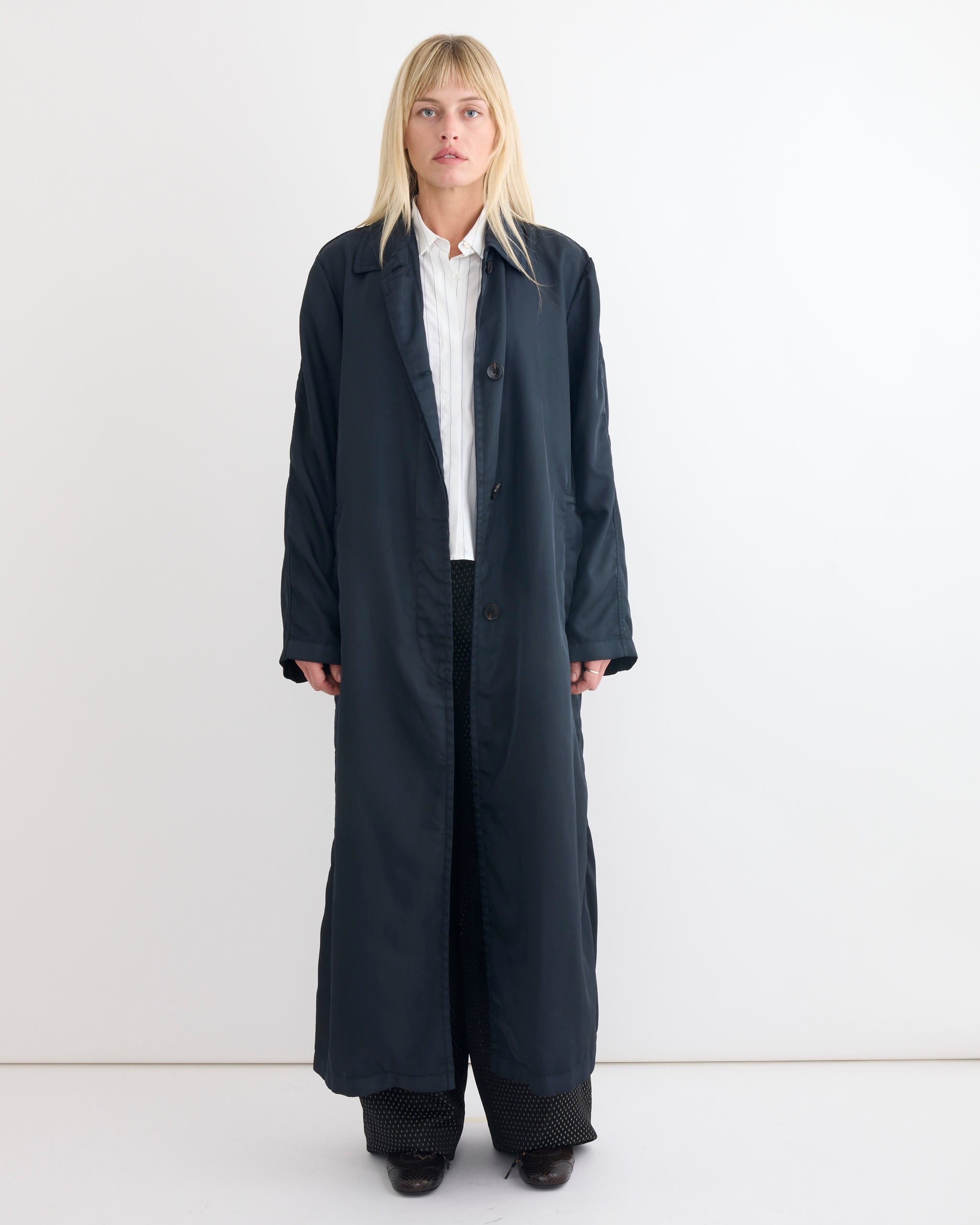 A person with straight blonde hair stands against a white background, wearing the Dries Van Noten Raltonas Coat in dark green over a white collared shirt and black pants.