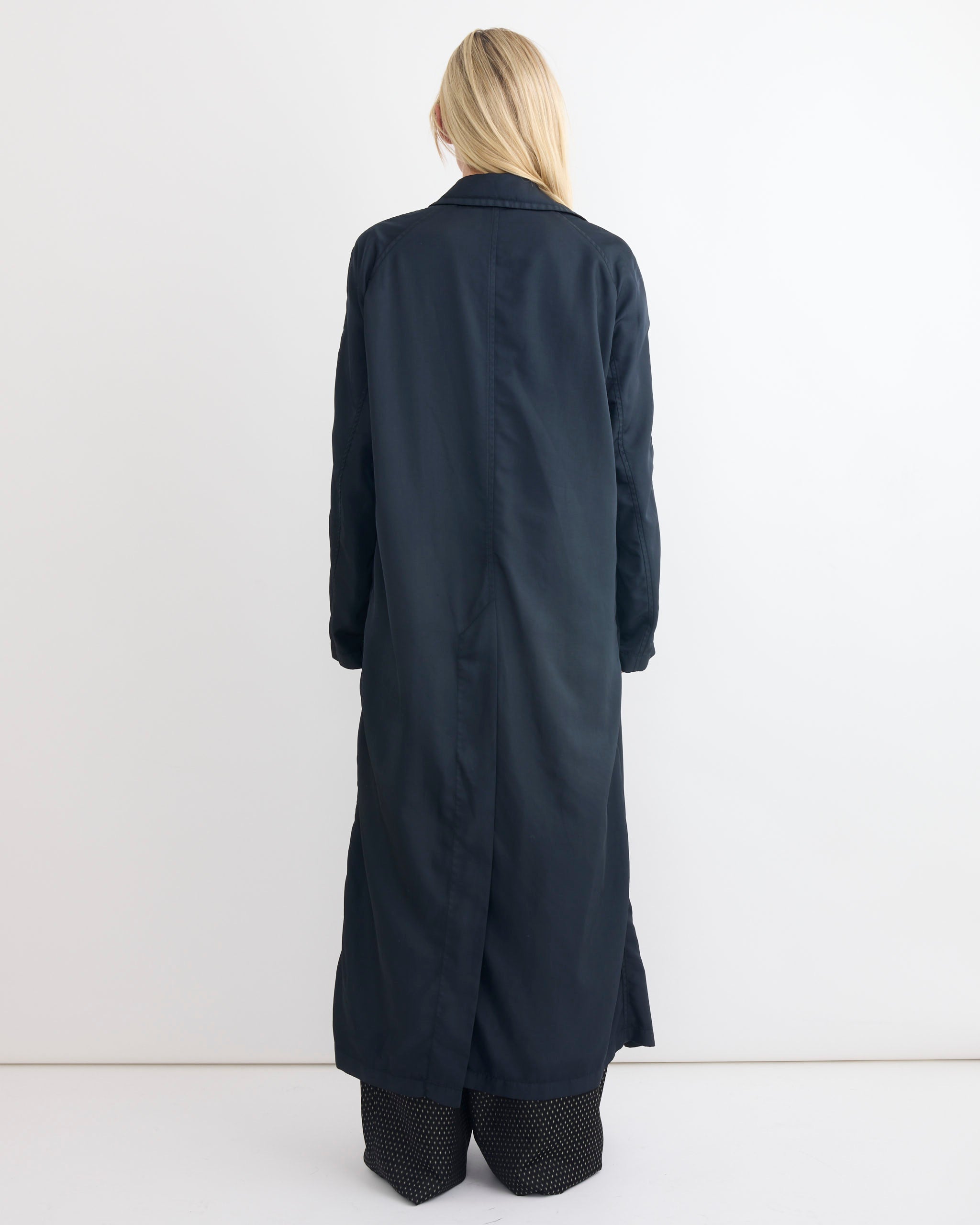 A person with long blonde hair stands facing away, wearing a dark green Dries Van Noten Raltonas Coat over black and white patterned pants against a plain white background.