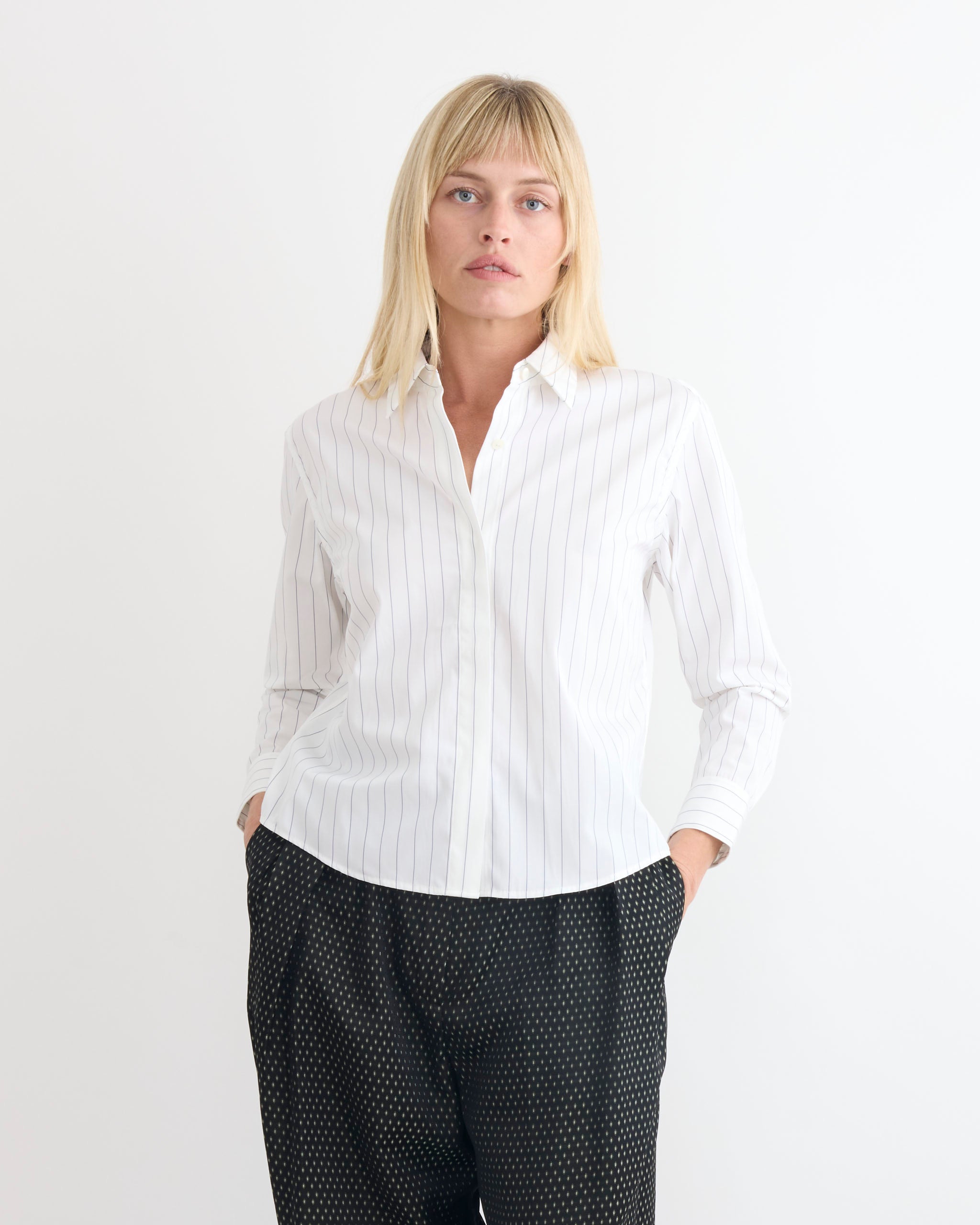 A person with straight blonde hair wears a Dries Van Noten Boxy Shirt in Navy and dark patterned pants, standing against a plain white background with hands in their pockets.