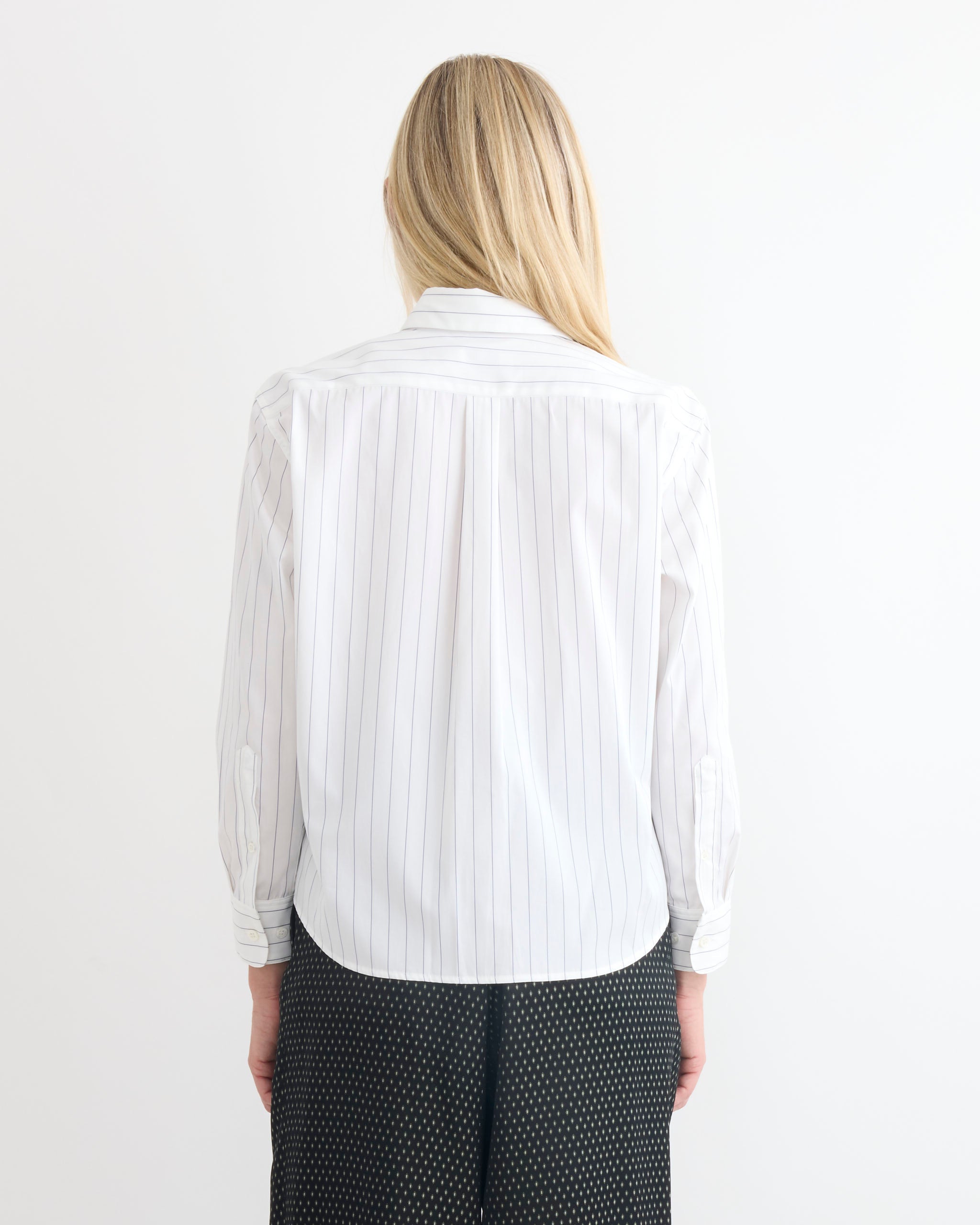 A person with long blonde hair stands facing away from the camera, wearing a Dries Van Noten Boxy Shirt in Navy and dark patterned pants, set against a plain white background.