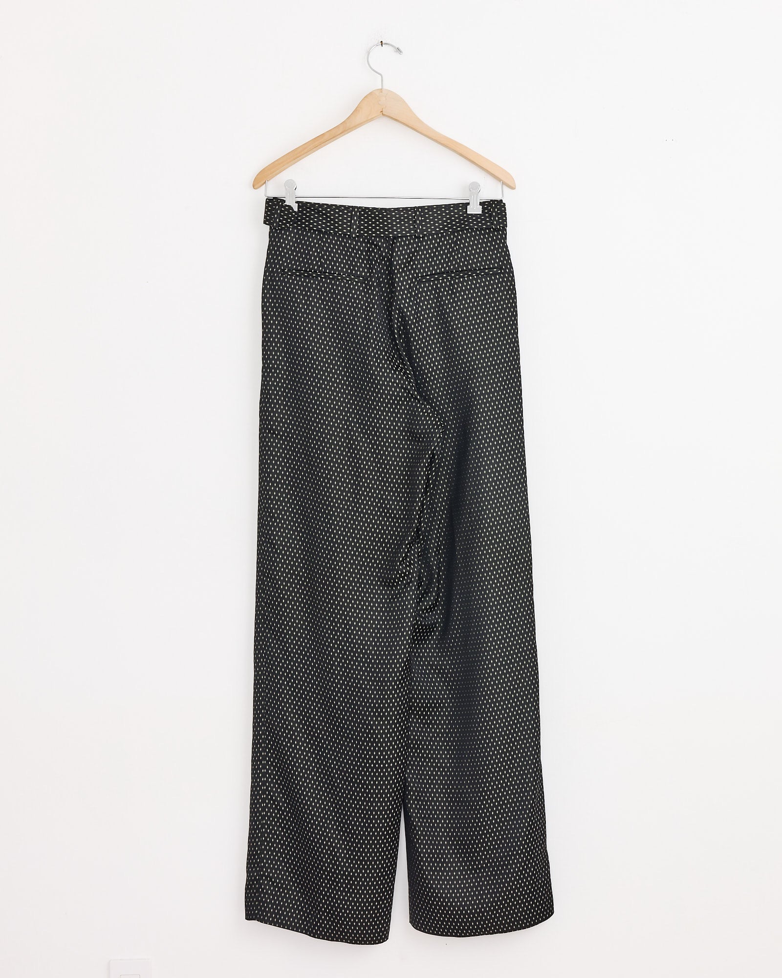 A pair of Dries Van Noten Belted Jacquard Trousers in Black, featuring a high-waisted, wide-leg silhouette and small white polka dots, hangs on a wooden hanger against a plain white wall.