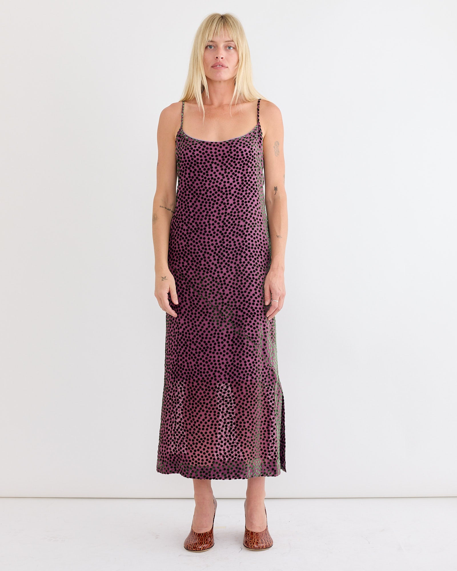 A woman with blonde hair stands against a white wall, wearing a sleeveless, ankle-length Dries Van Noten Slip Dress in Dark Pink with thin straps. She faces forward with a neutral expression.