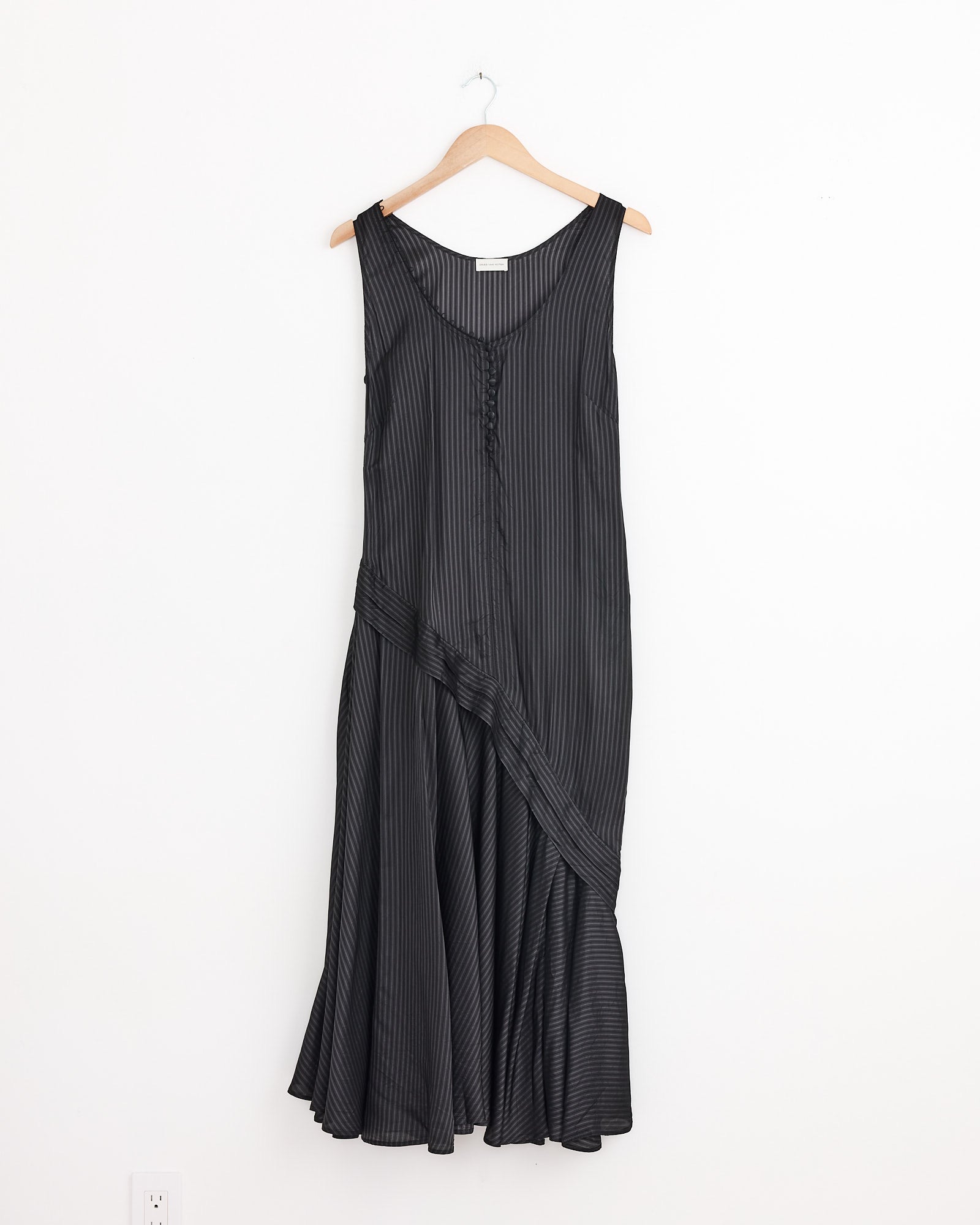 The Dommy Stripe Crepe Midi Dress in Grey by Dries Van Noten, with thin vertical stripes, a V-neckline, and an asymmetric draped hem, hangs on a wooden hanger against a plain white wall.