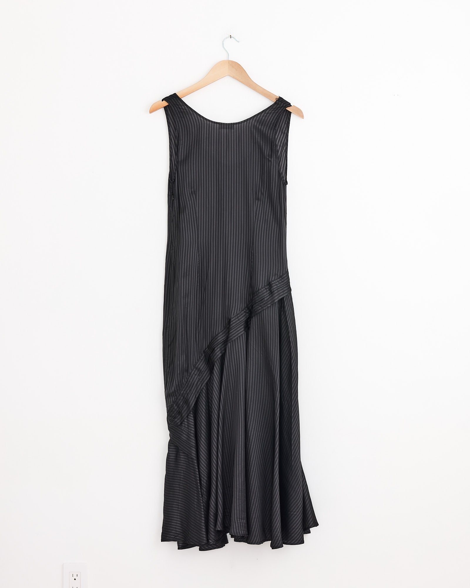 A sleeveless, ankle-length grey Dries Van Noten Dommy Stripe Crepe Midi Dress with subtle vertical stripes and asymmetric draping across the skirt, displayed on a wooden hanger against a white wall.