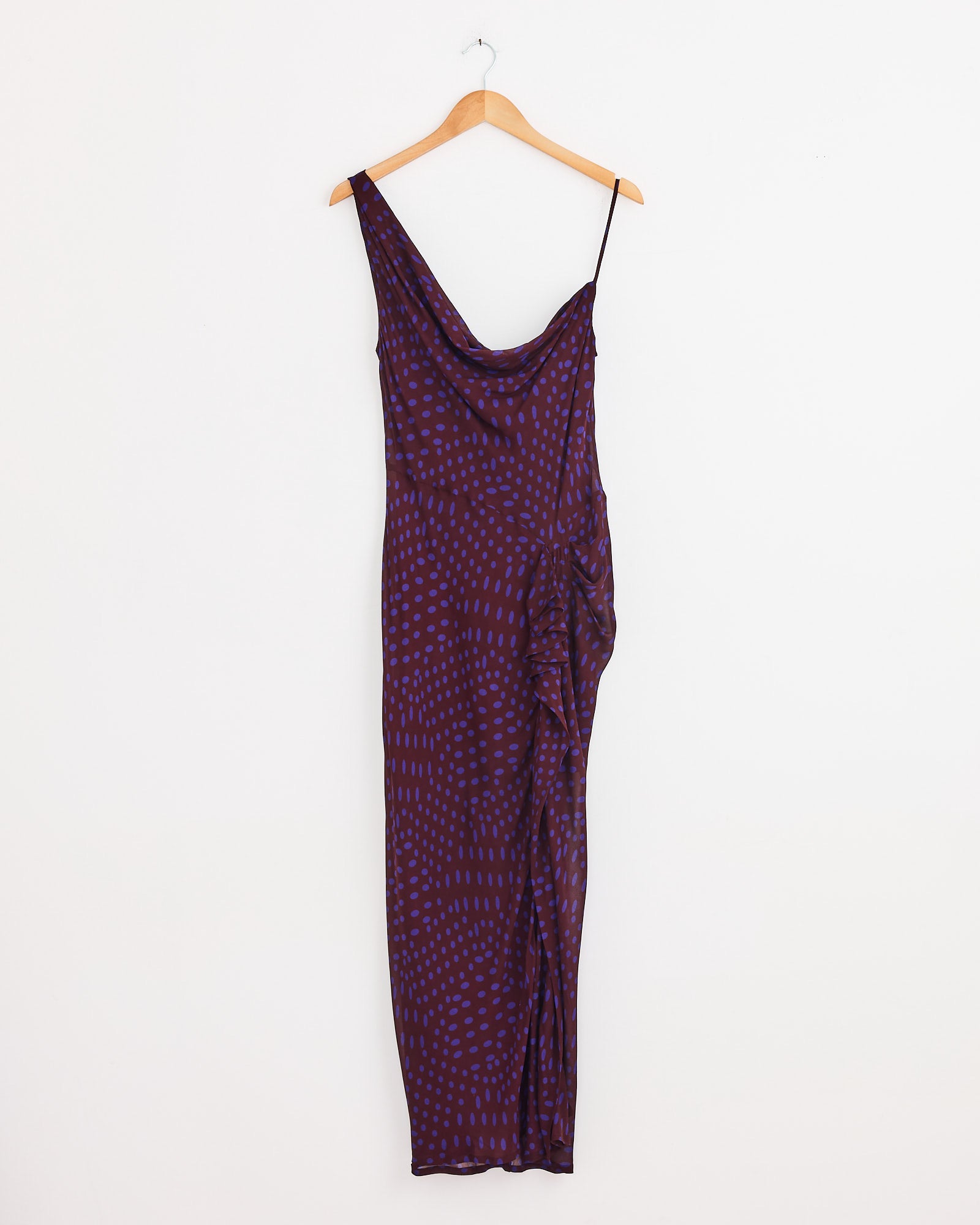 The Dries Van Noten Asymmetric Dress in Auber, a long sleeveless viscose georgette style with thin straps and a draped neckline, features a dark purple base with small red shapes and hangs on a wooden hanger against a white wall.