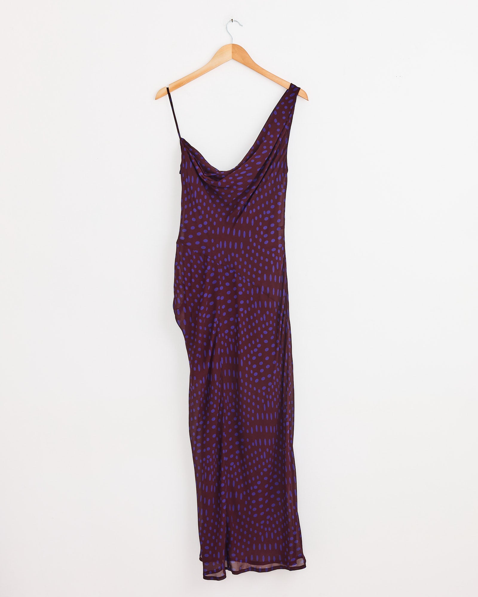 The Asymmetric Dress in Auber by Dries Van Noten, a long sleeveless design with thin straps and a subtle purple dotted print, hangs on a wooden hanger against a plain white wall.