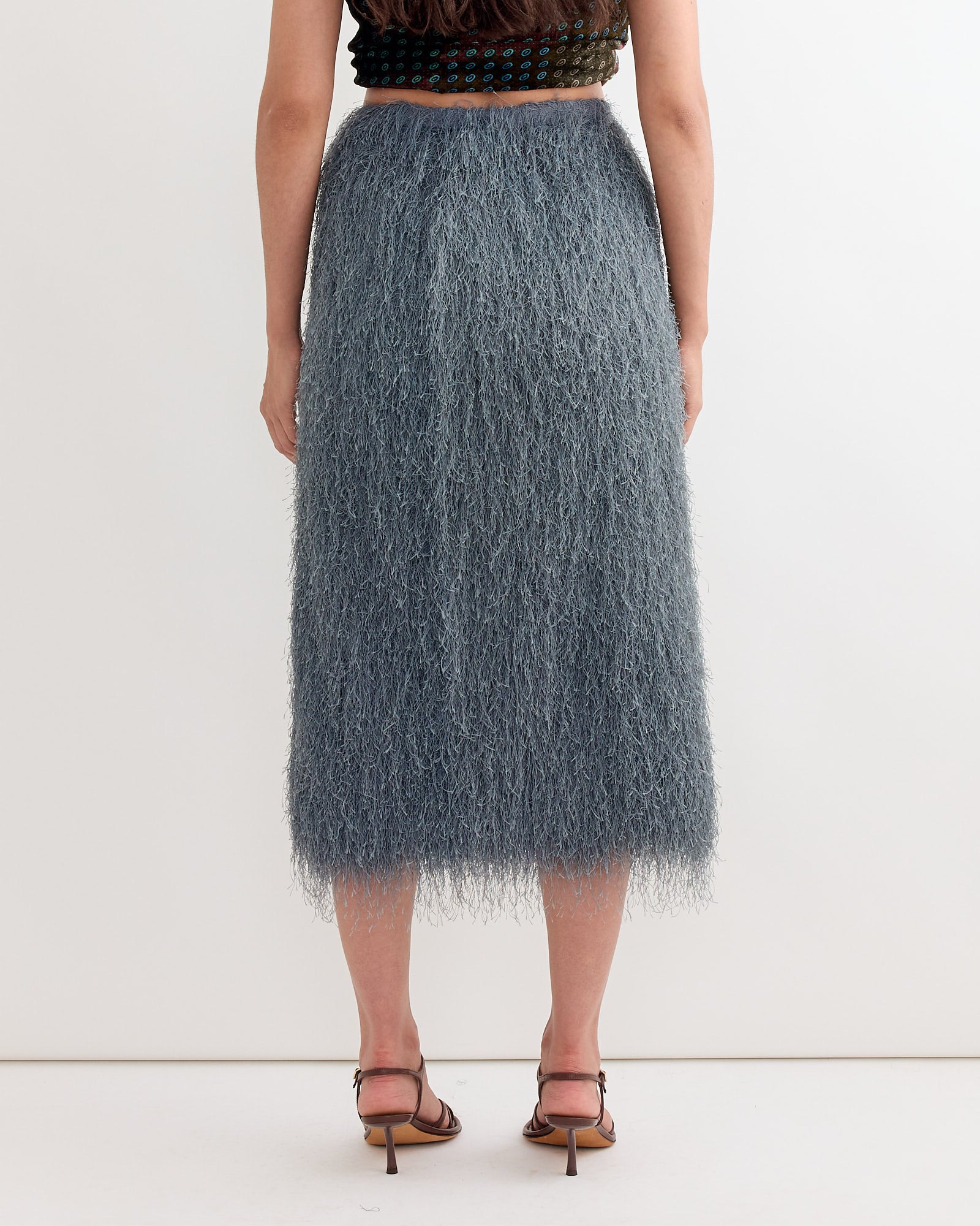 A person stands facing away, wearing a Dries Van Noten Fringed Skirt in Dark Grey, paired with strappy high-heeled sandals and a black sleeveless top.