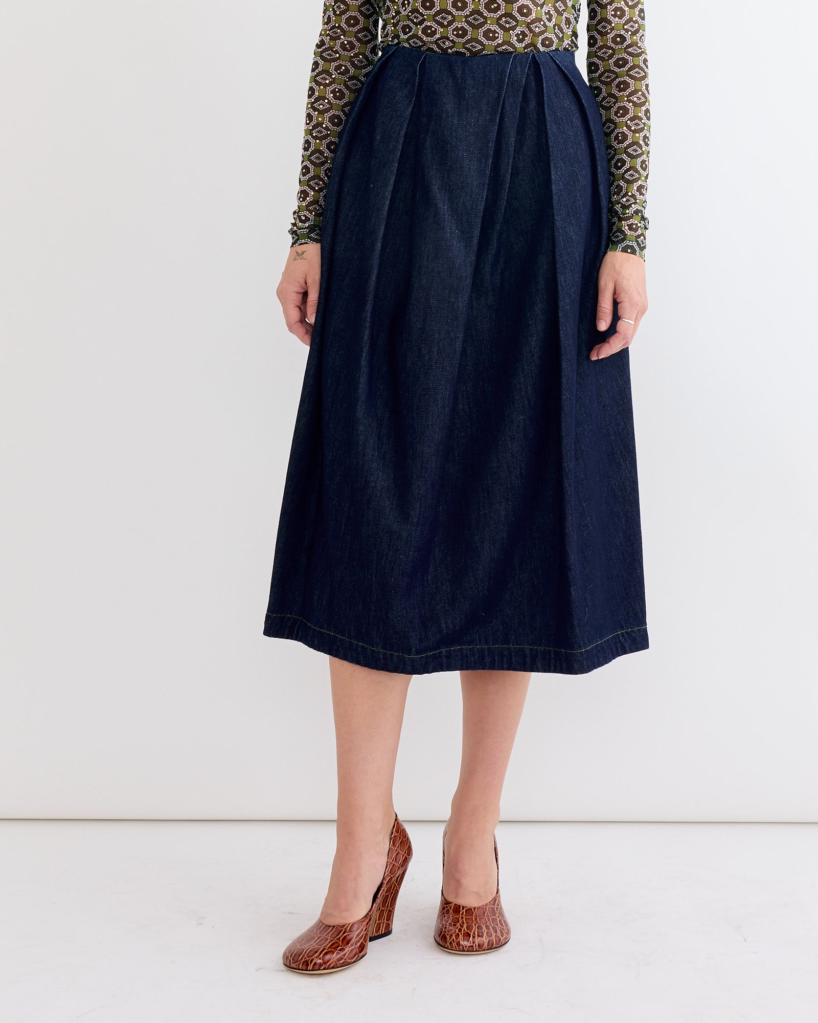 Woman in a patterned long-sleeve top, Dries Van Noten Pleated Denim Skirt in Indigo, and brown textured heels, standing against a plain white background.