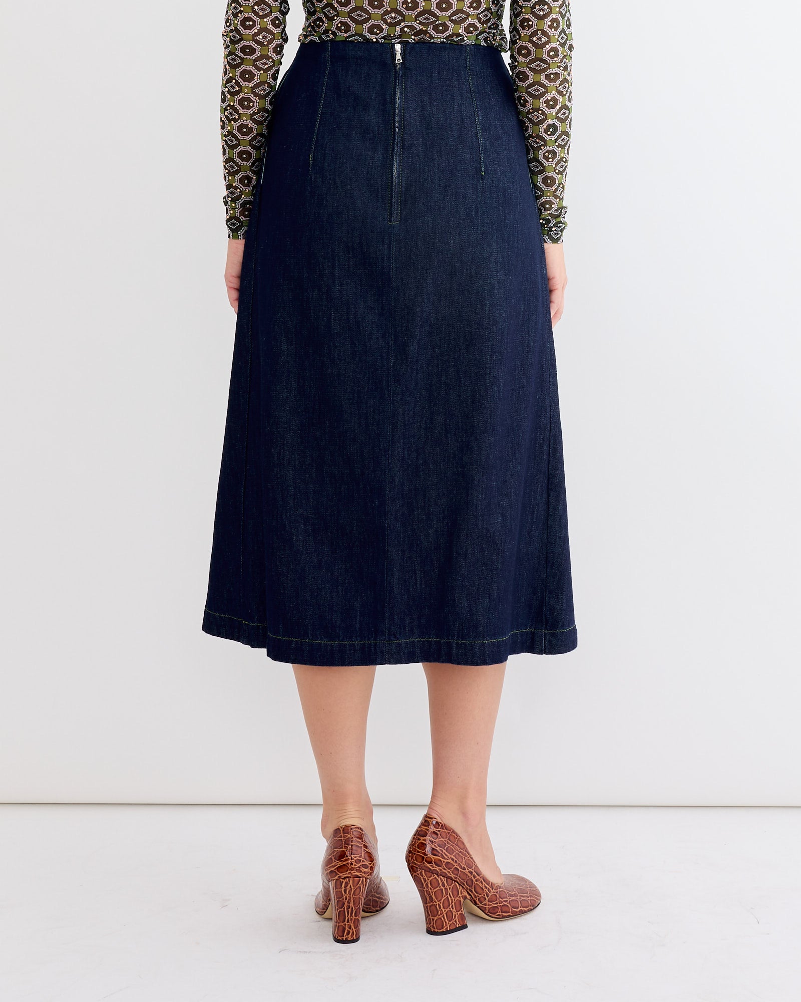 A person faces away, wearing a patterned long-sleeve top, the Pleated Denim Skirt in Indigo by Dries Van Noten with a visible back zipper, and brown textured block heels, set against a plain white background.