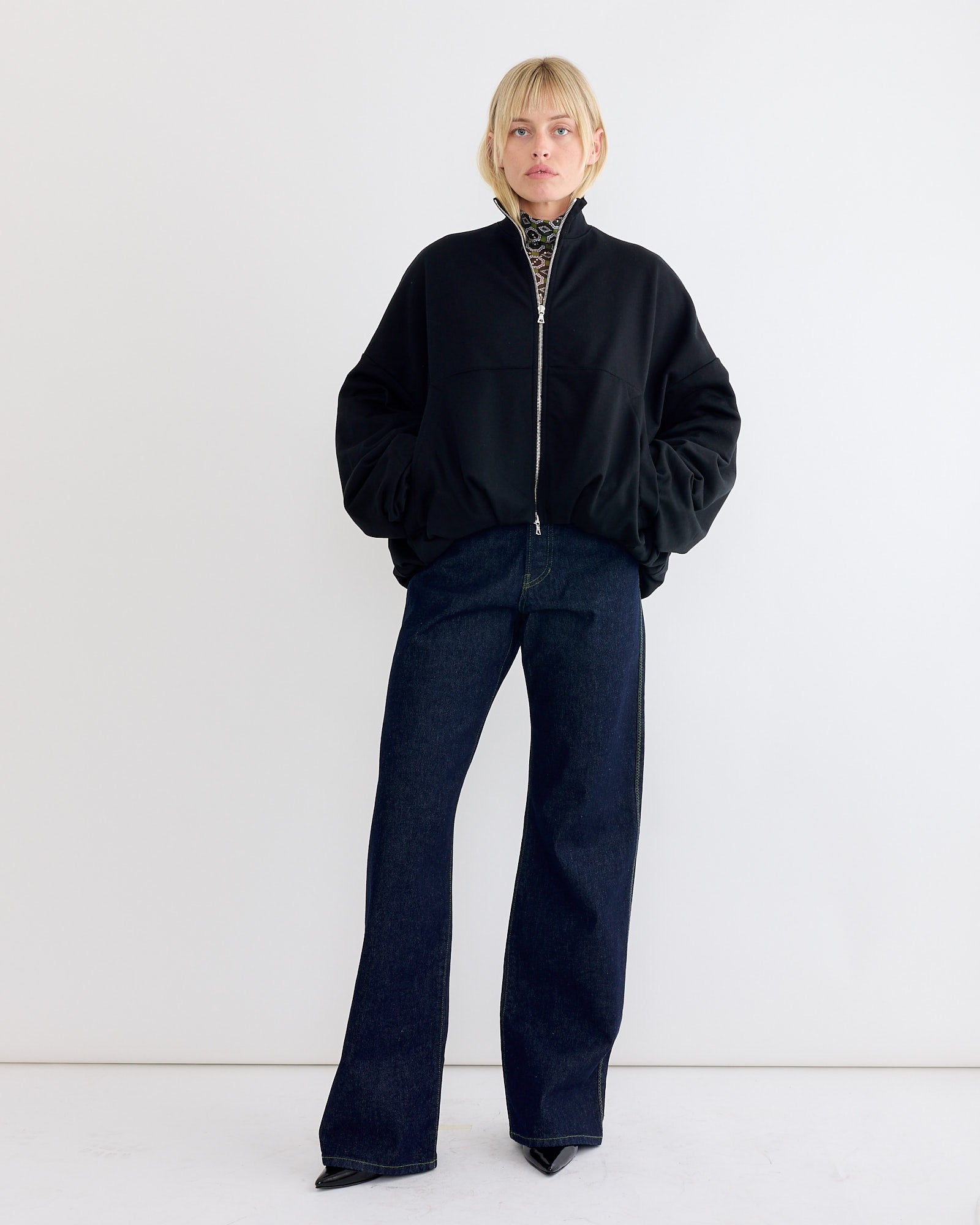 A person stands against a white background wearing an oversized silhouette featuring the Dries Van Noten Draped Jersey Jacket in Black, dark wide-leg jeans, pointed black shoes, and a patterned high-neck top, with hands in the jacket pockets.