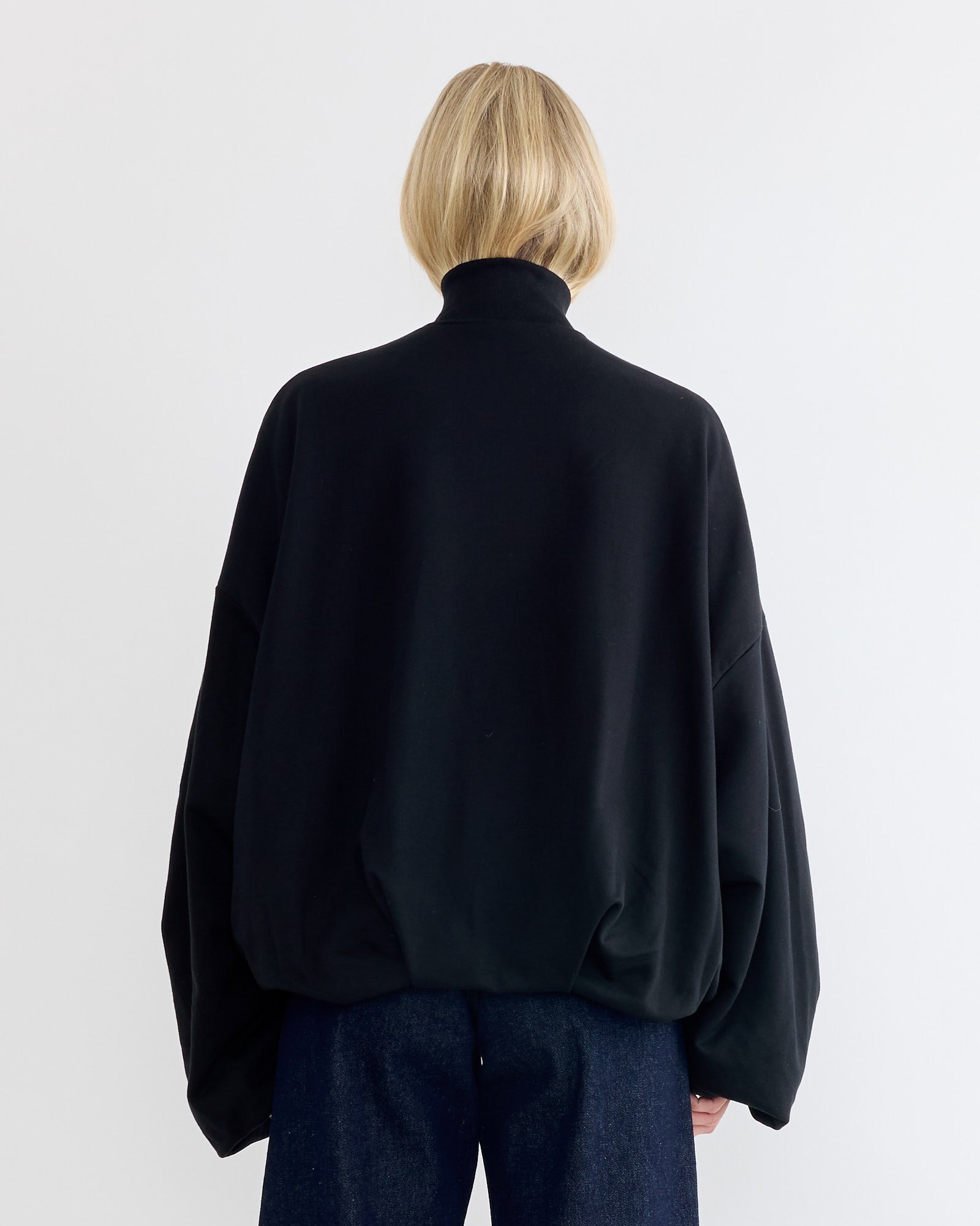 A person with blonde hair, seen from behind, wears a Dries Van Noten Draped Jersey Jacket in black and dark blue jeans, standing against a plain white background.