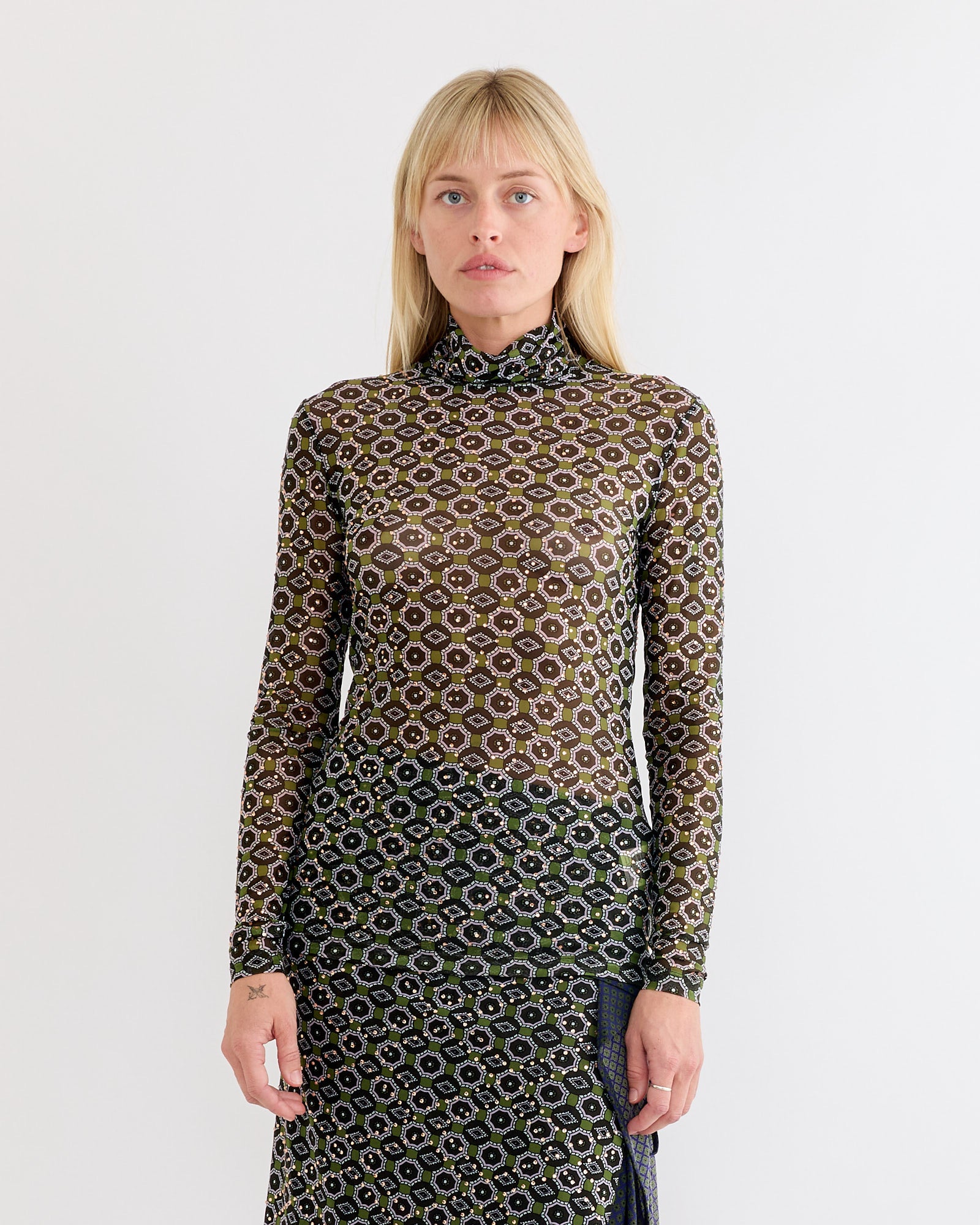 A person with straight blonde hair and bangs stands against a white background, wearing a Dries Van Noten Embellished Turtleneck Tee in Green.