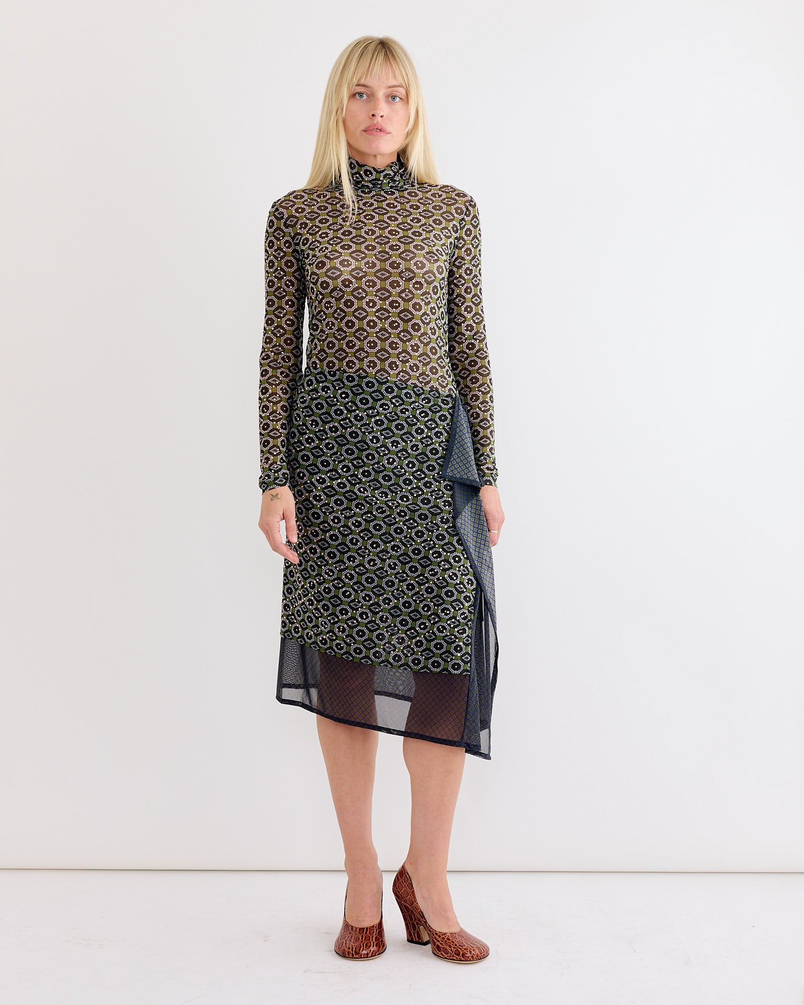 A woman stands against a white background wearing a long-sleeve, high-neck, geometric-patterned top and the Dries Van Noten Embellished Skirt in Green. She has straight blonde hair and brown textured heels.