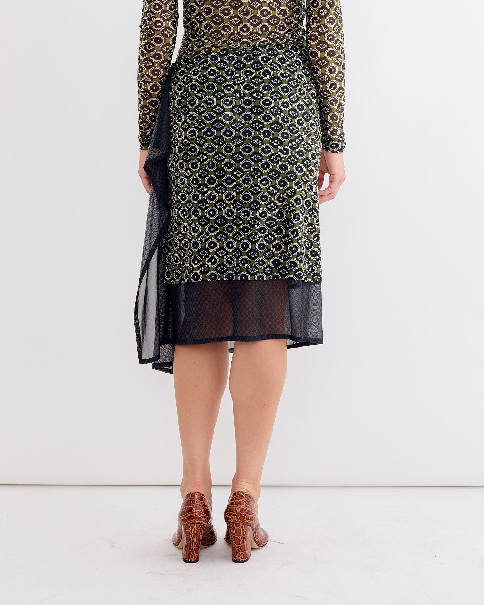 A person stands facing away, wearing a Dries Van Noten Embellished Skirt in Green with sheer, patterned layers over a black slip and brown heeled shoes, set against a plain white background.