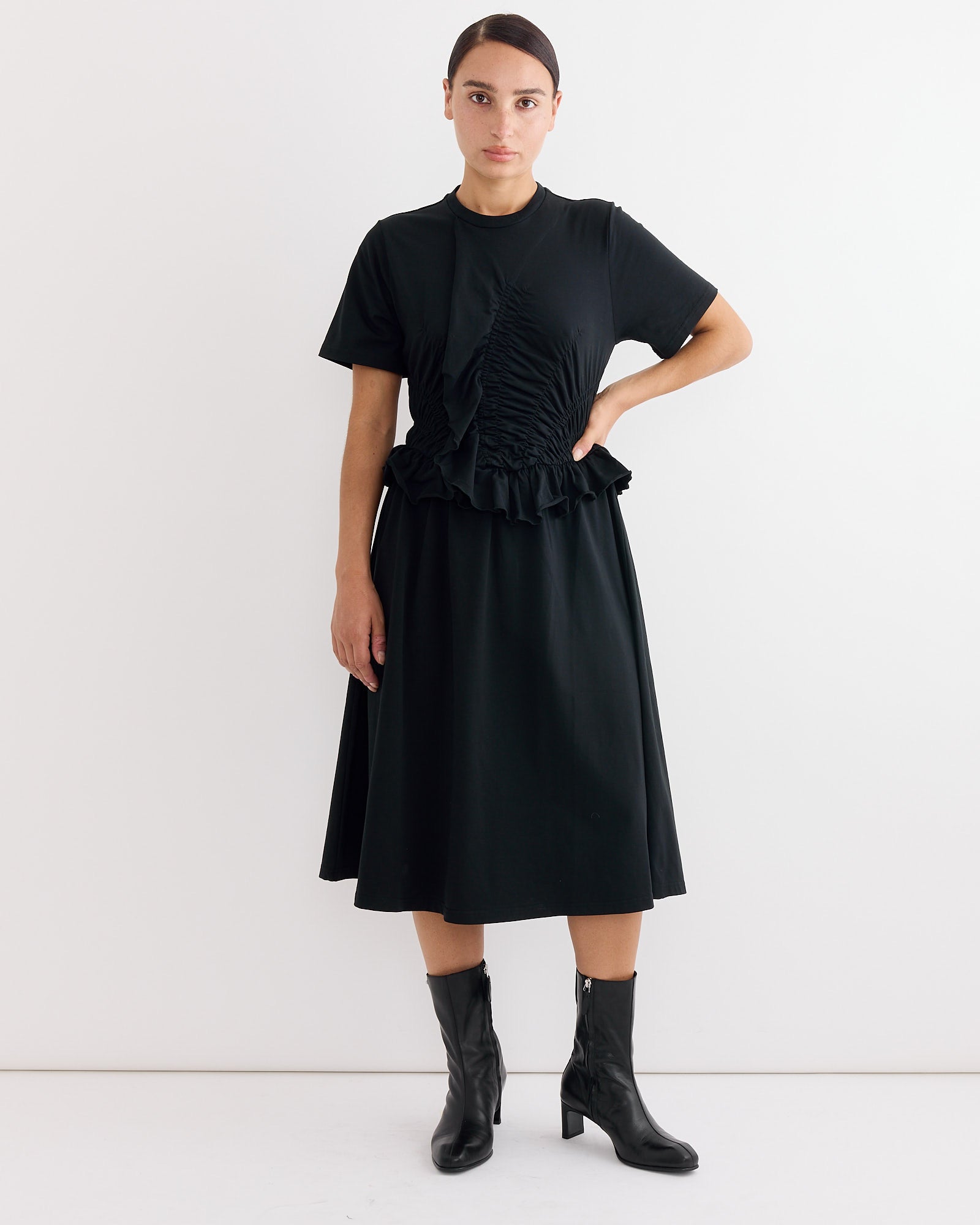 A person stands against a plain white background wearing the Cecilie Bahnsen Bonnie Dress in Black, styled with black heeled boots. One hand rests on their hip, and they have a neutral expression.