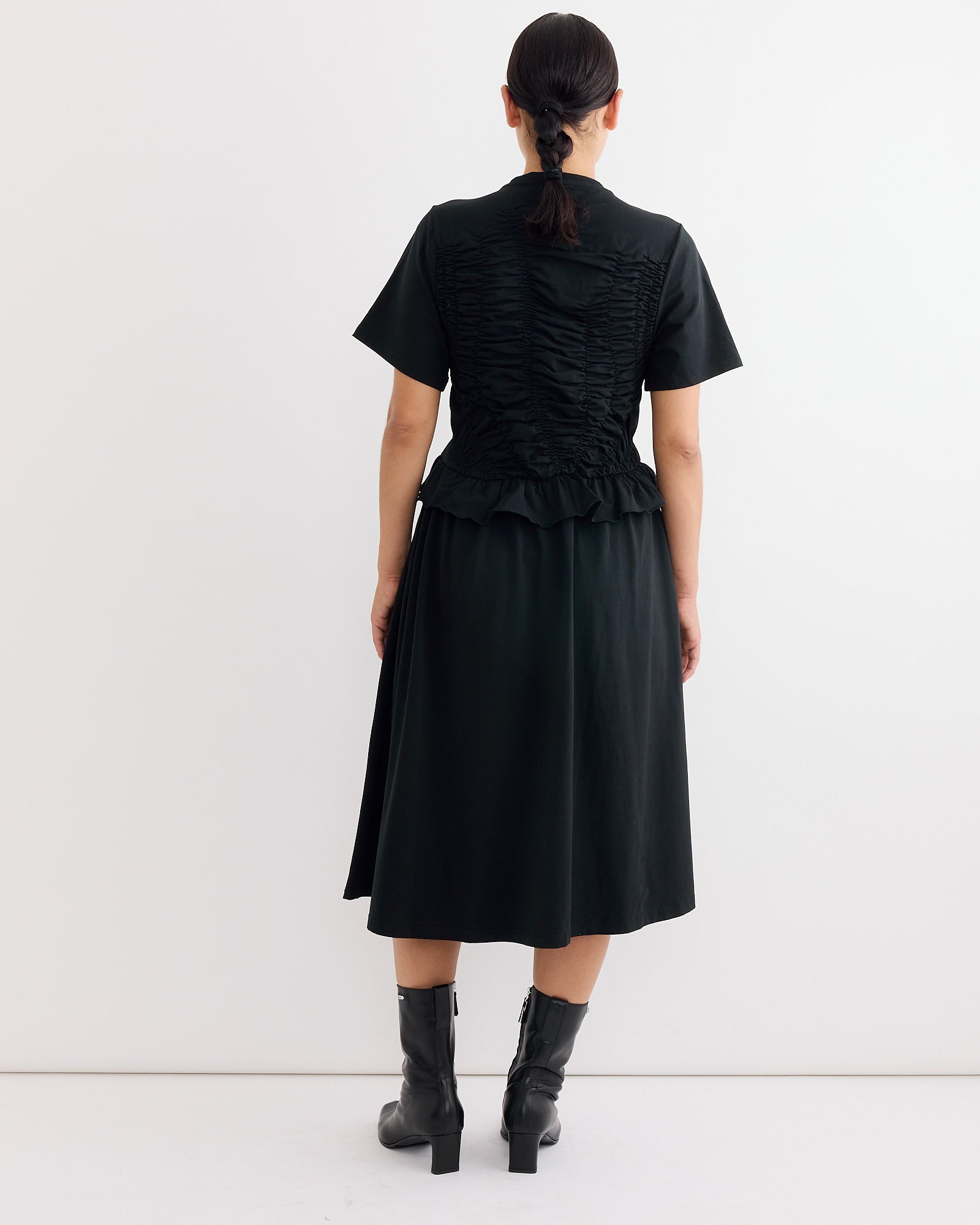 A person stands facing away, wearing the Cecilie Bahnsen Bonnie Dress in Black—a t-shirt dress with short sleeves and a ruched back—paired with black mid-calf boots. Their hair is tied back against a plain white background.