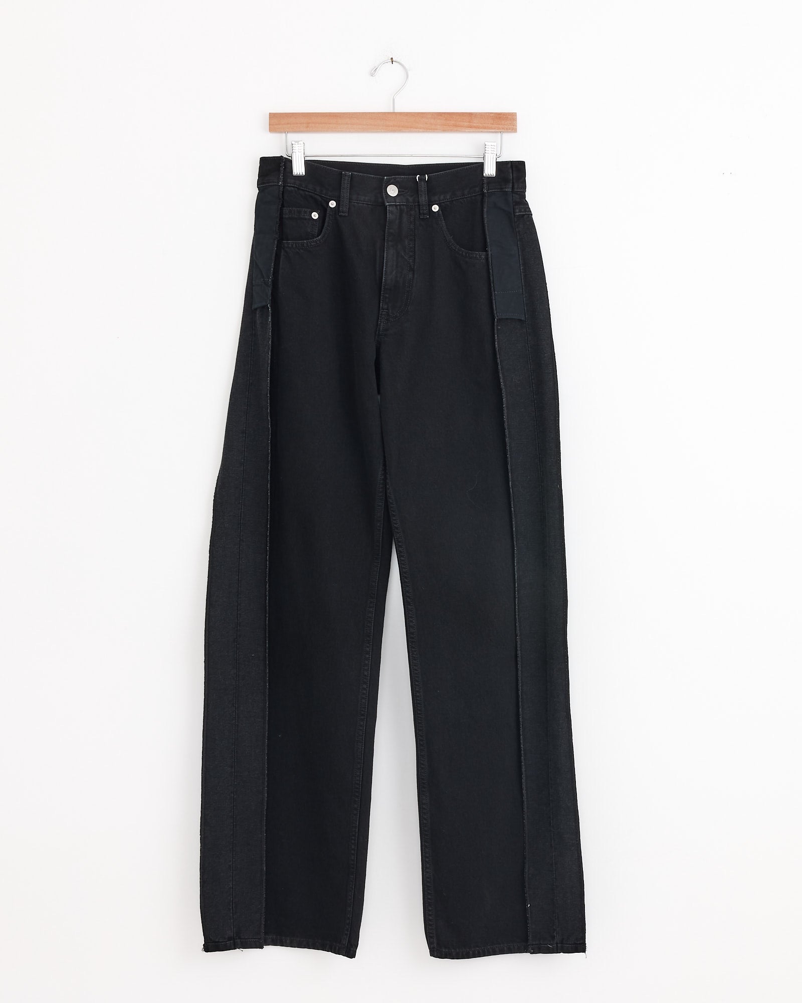 The MM6 Maison Margiela 5 Pockets Pant in Black features relaxed wide-leg cotton denim with sculptural detailing and contrasting side panels, hanging on a wooden hanger against a white background for a modern look.