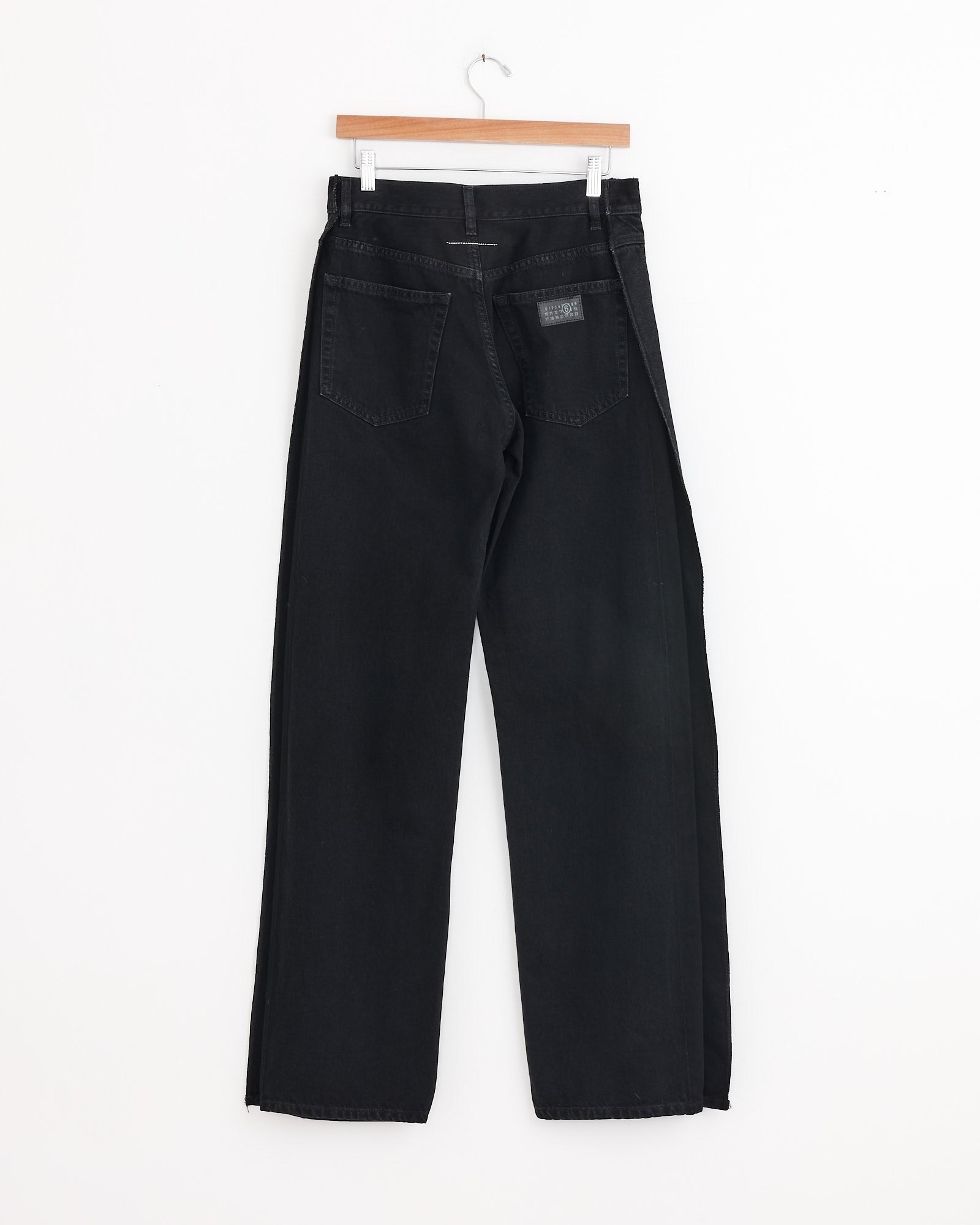 The MM6 Maison Margiela 5 Pockets Pant in Black, crafted from cotton denim with a straight-leg fit, hangs on a wooden hanger with the back facing forward against a plain white background for a sculptural effect.