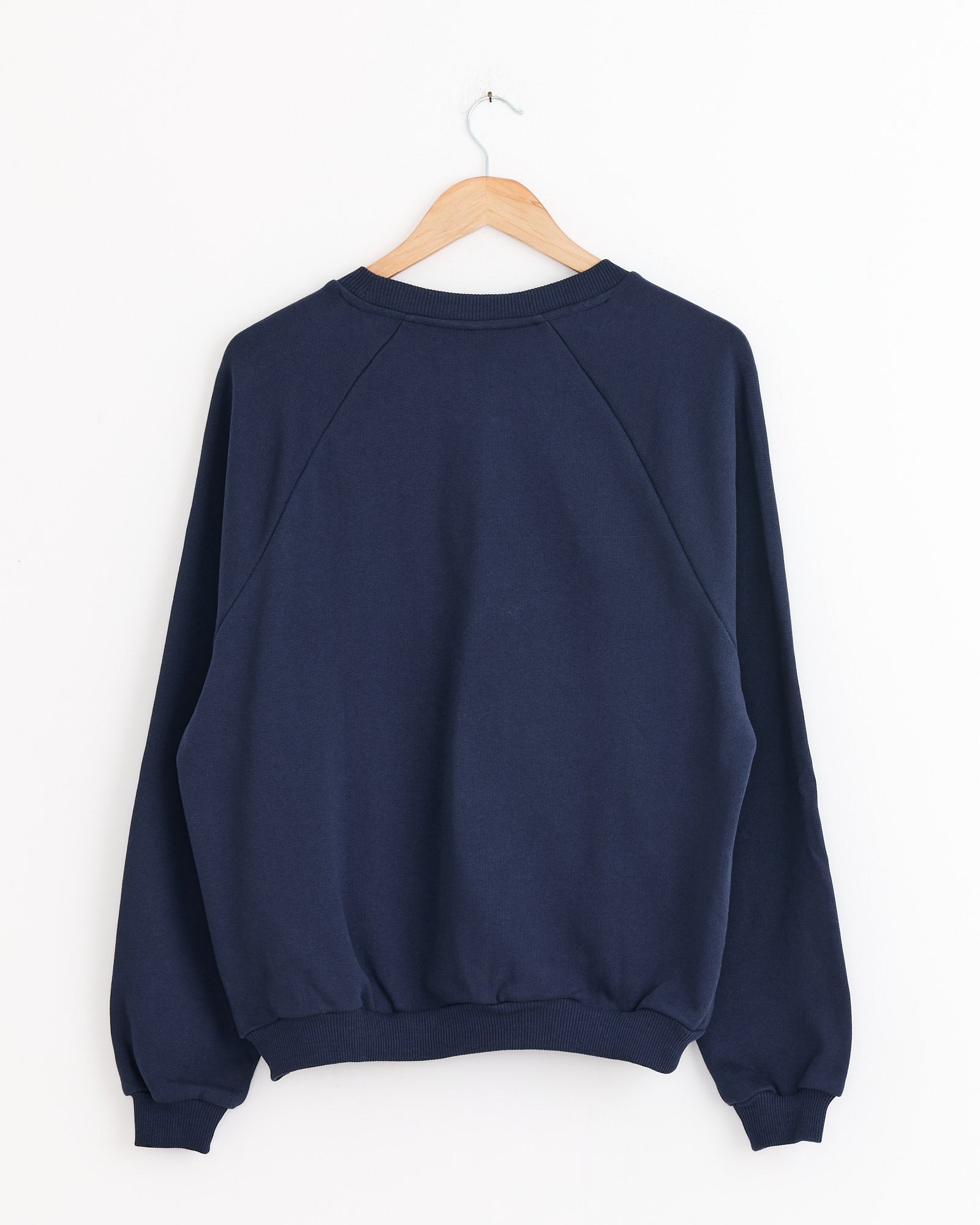 The Baserange Rim Raglan Sweatshirt in Echo Blue, made from soft organic cotton with long sleeves and ribbed cuffs and hem, is shown hanging on a wooden hanger against a white wall, displayed from the back.
