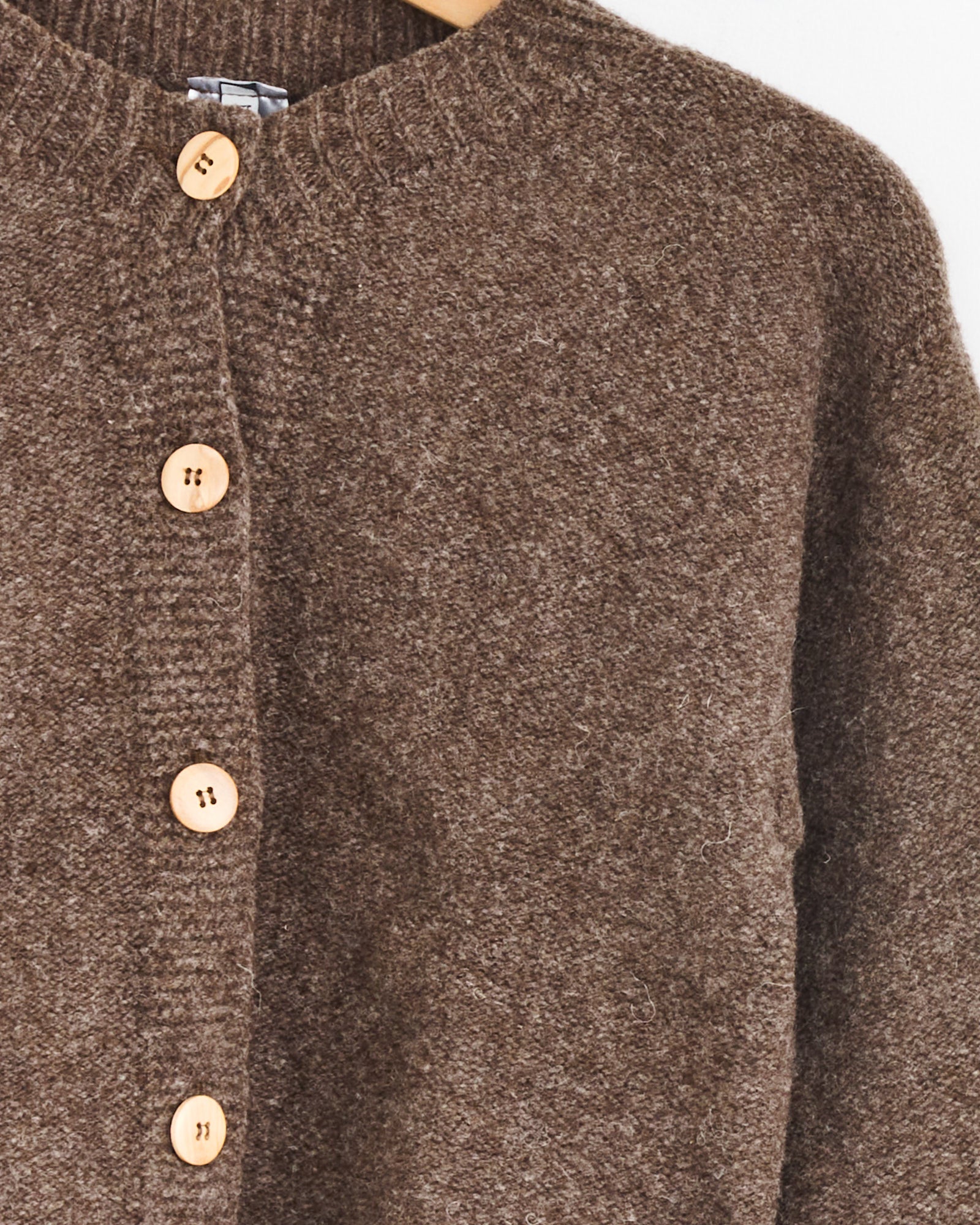 A close-up of the Baserange Tier Cardigan in Burel Brown, featuring light wooden buttons and a round neckline, hanging on a hanger against a plain white background.