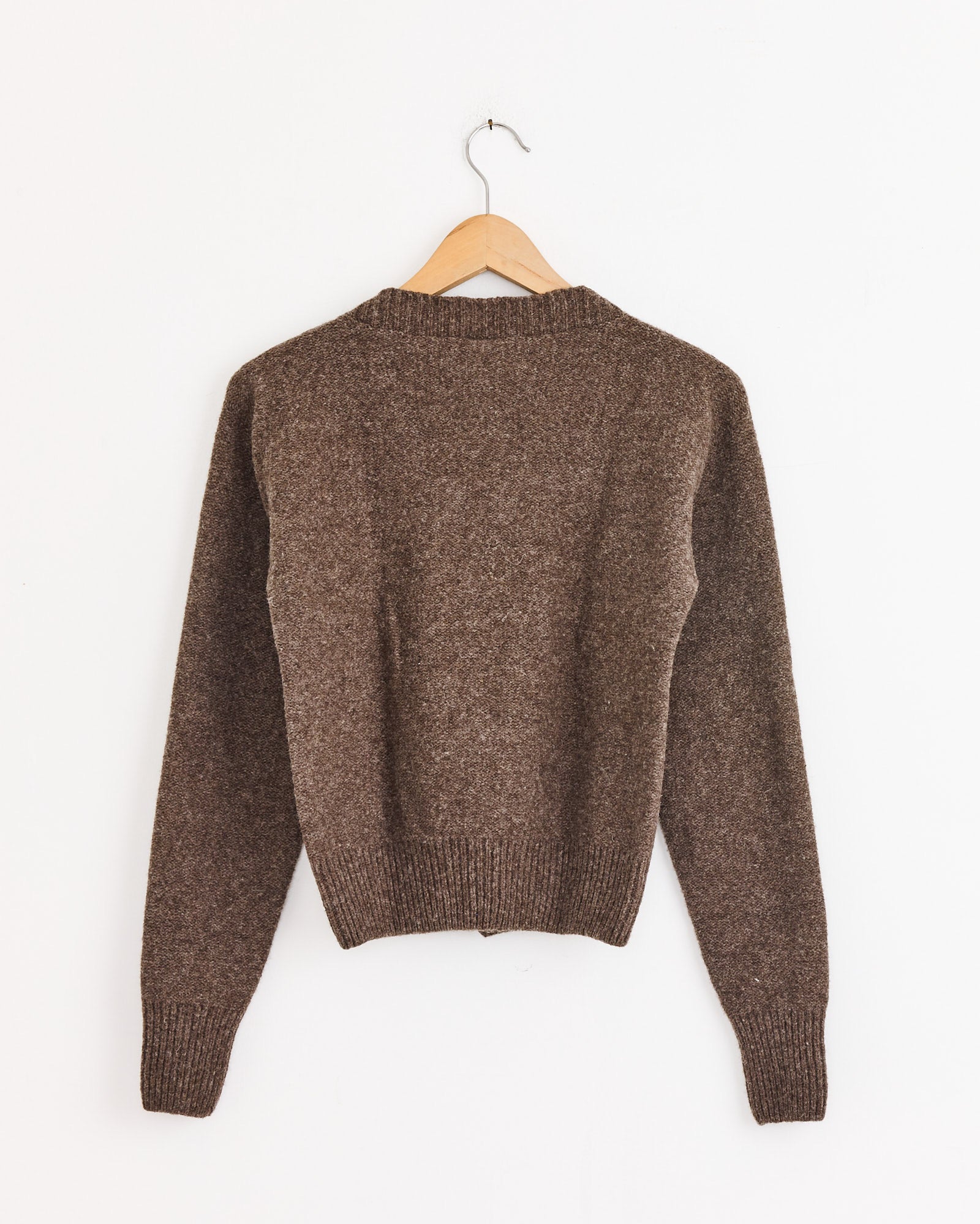 The Baserange Tier Cardigan in Burel Brown, made from Arles merino wool with long sleeves, is displayed on a wooden hanger against a plain white wall, shown from the back.
