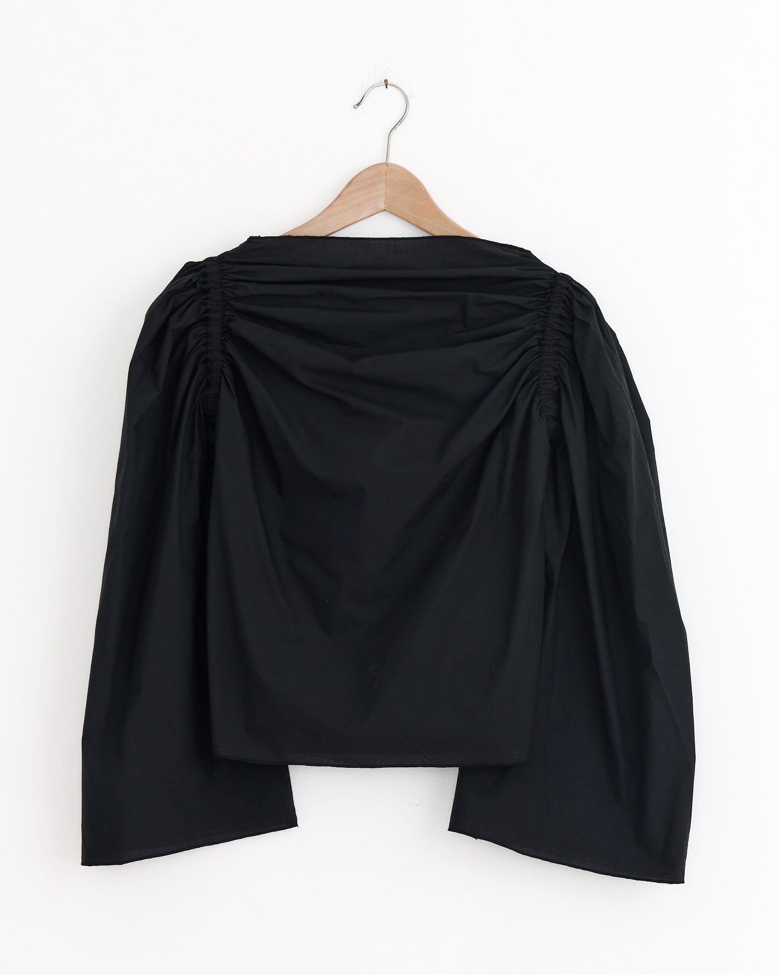 The Baserange Simmon Top in Black, a long-sleeve organic cotton poplin blouse with ruched shoulders, loose fit, and wide sleeves, is shown on a wooden hanger against a white background.