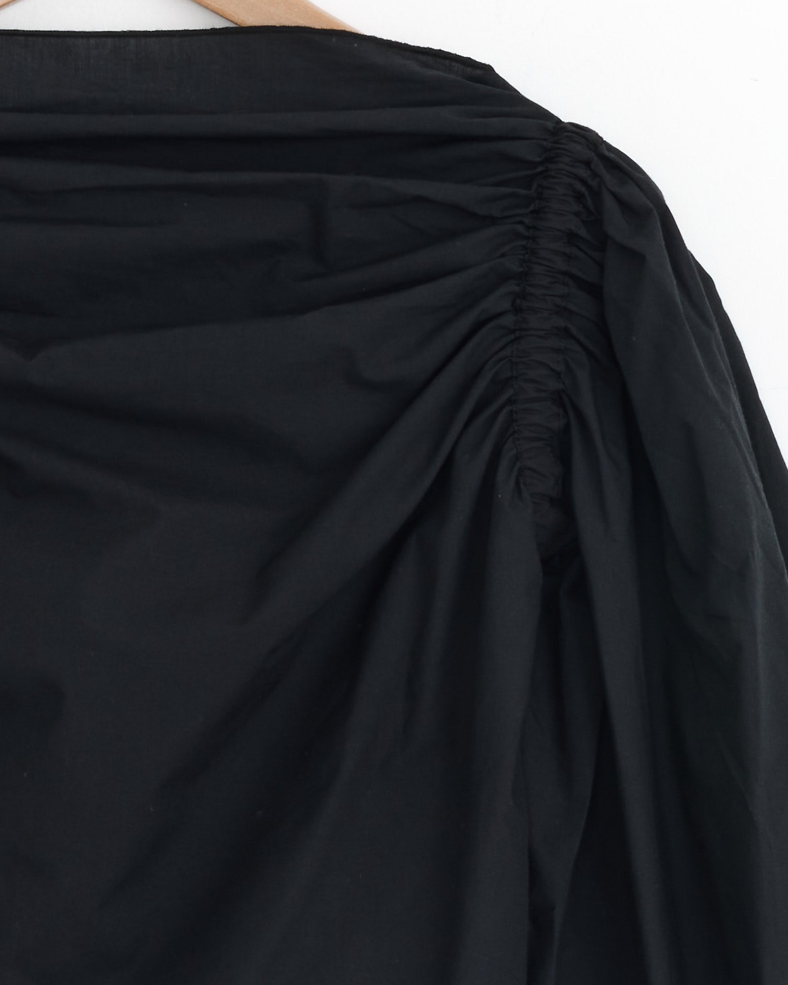 Close-up of the Baserange Simmon Top in Black, with gathered ruched shoulder seams and smooth, slightly shiny organic cotton poplin, shown on a wooden hanger against a plain white background.