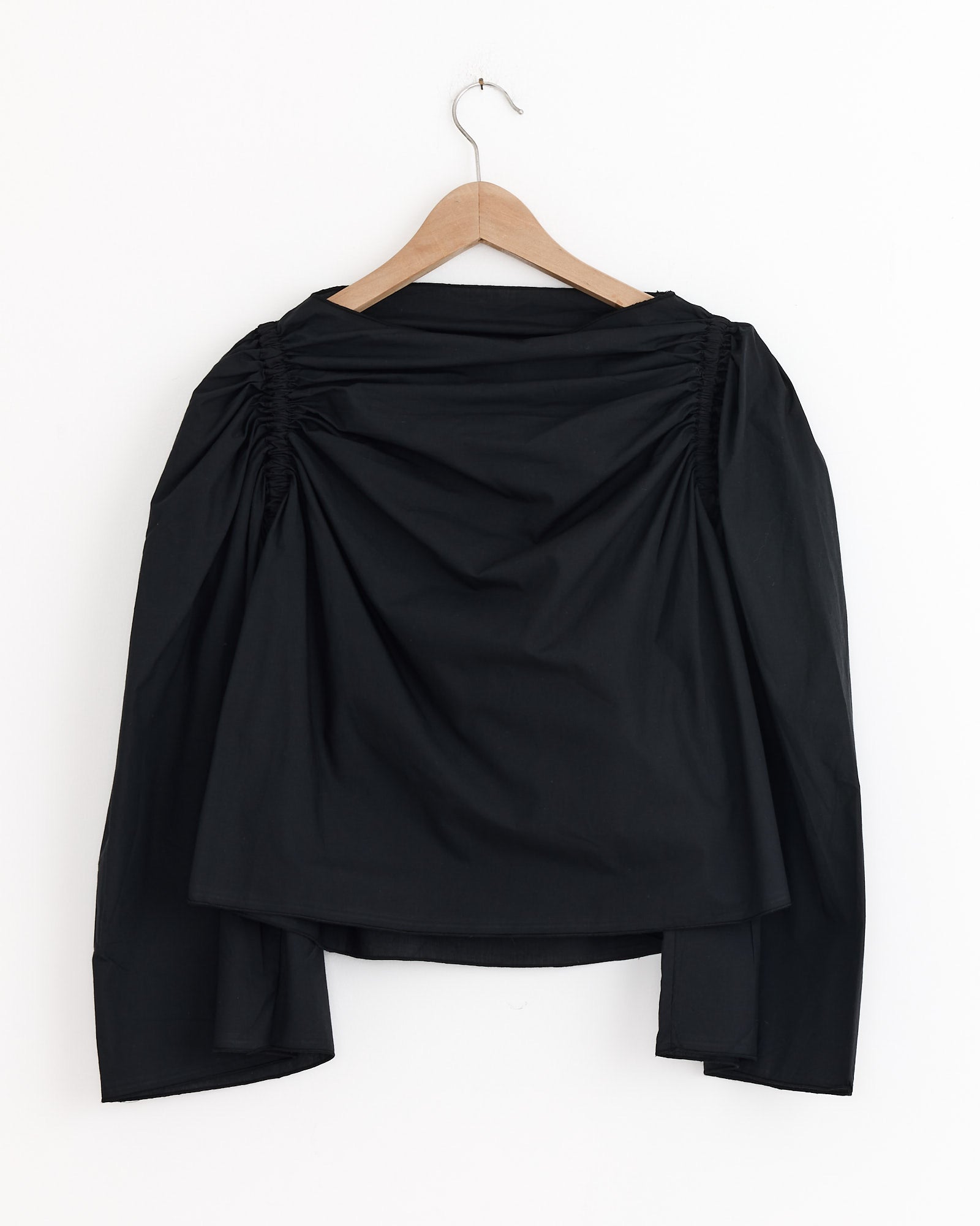 The Simmon Top in Black by Baserange is a long-sleeve blouse with ruched shoulder detailing, made from organic cotton poplin. It’s shown on a wooden hanger against a white background.