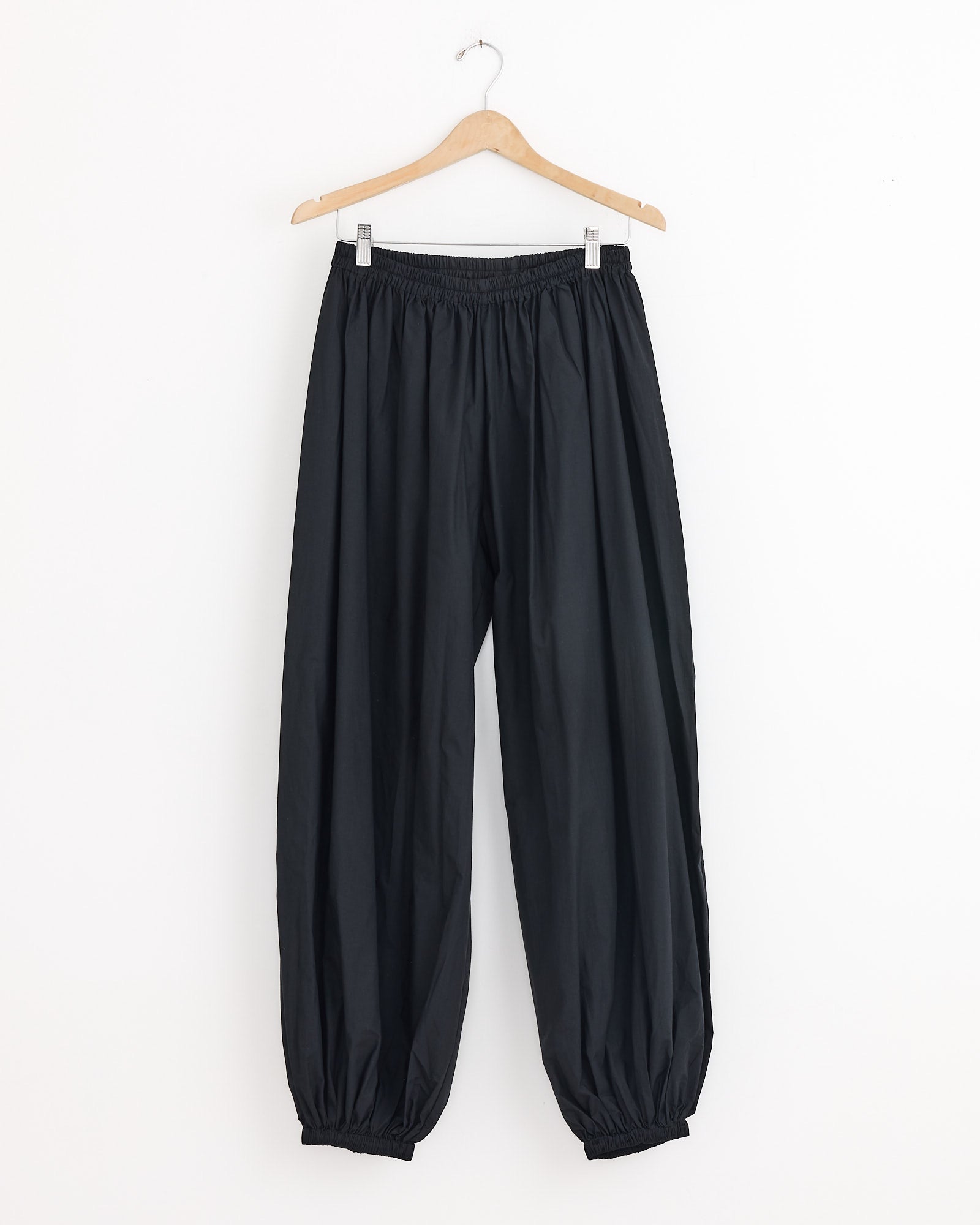 The Simmon Pant in Black by Baserange features a relaxed fit, lightweight breathable fabric, and elastic waistband and cuffs, shown hanging on a wooden hanger against a plain white background.