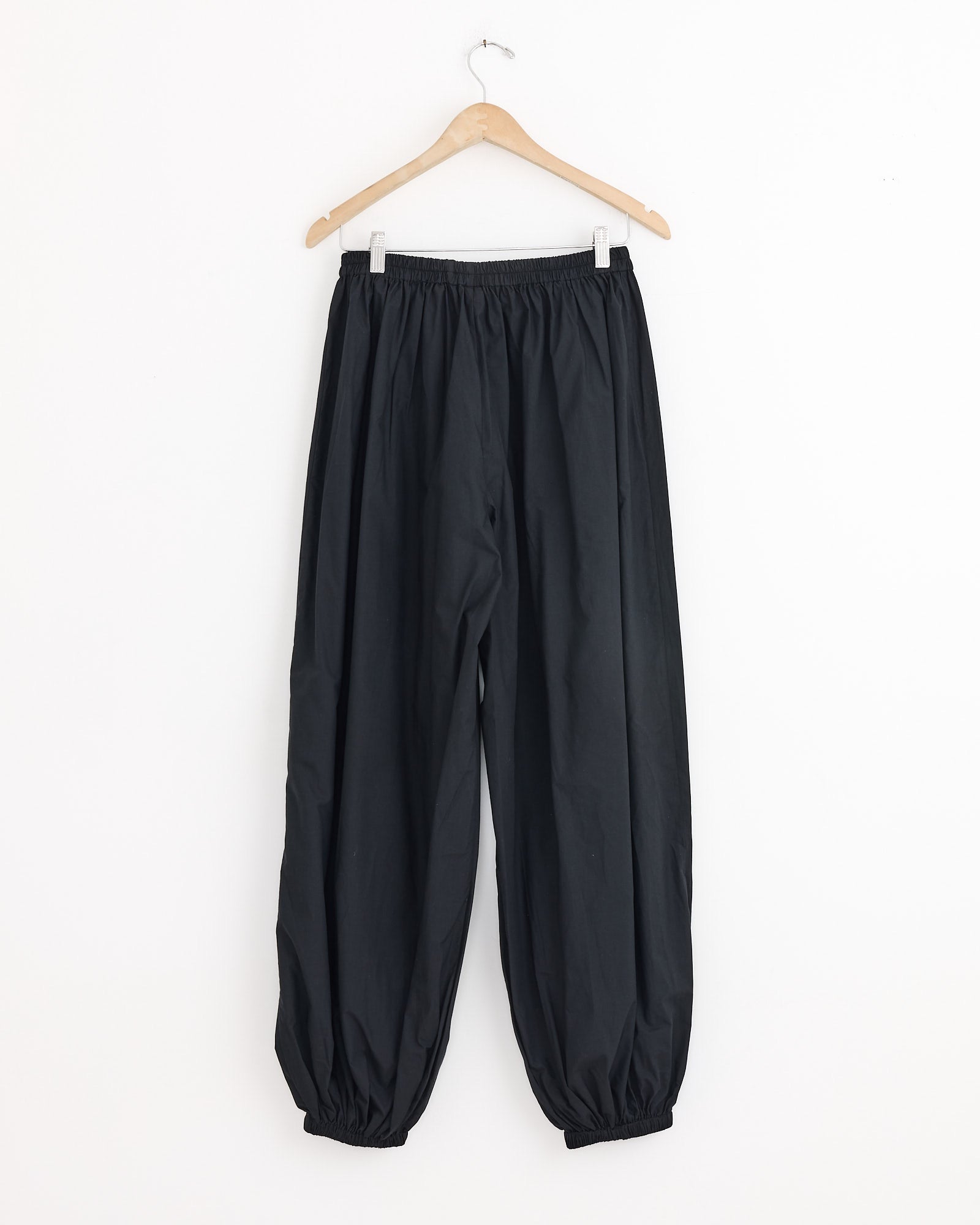 The Simmon Pant in Black by Baserange is a relaxed-fit, organic cotton pant with elastic at the waist and ankles, shown on a wooden hanger against a plain white background.