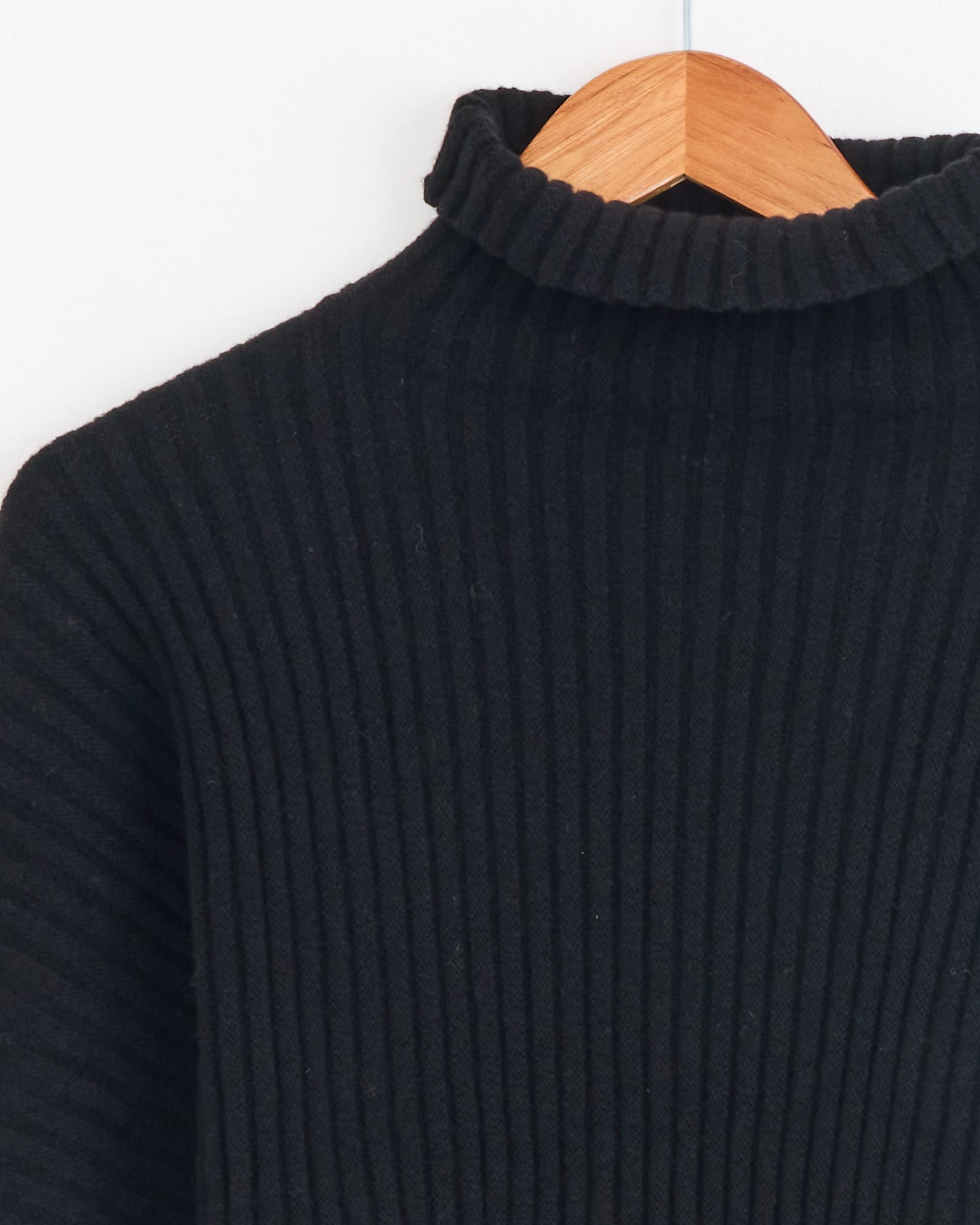Close-up of the Baserange Kai Turtleneck in Black on a wooden hanger, highlighting the ribbed neckline and upper half—crafted from soft recycled cashmere—against a plain white background.